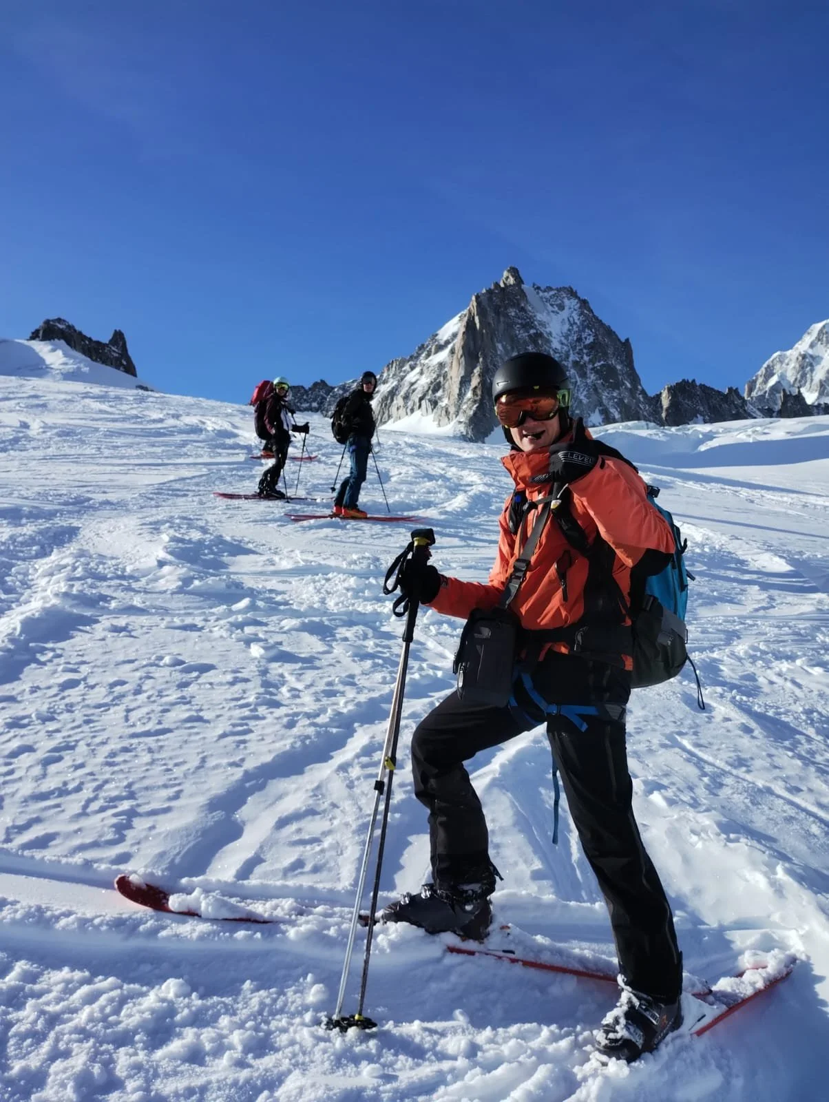 Group of four skiers on a snowy mountain with rocky peaks in the background, bright blue sky, smiling, wearing winter gear, and carrying backpacks.