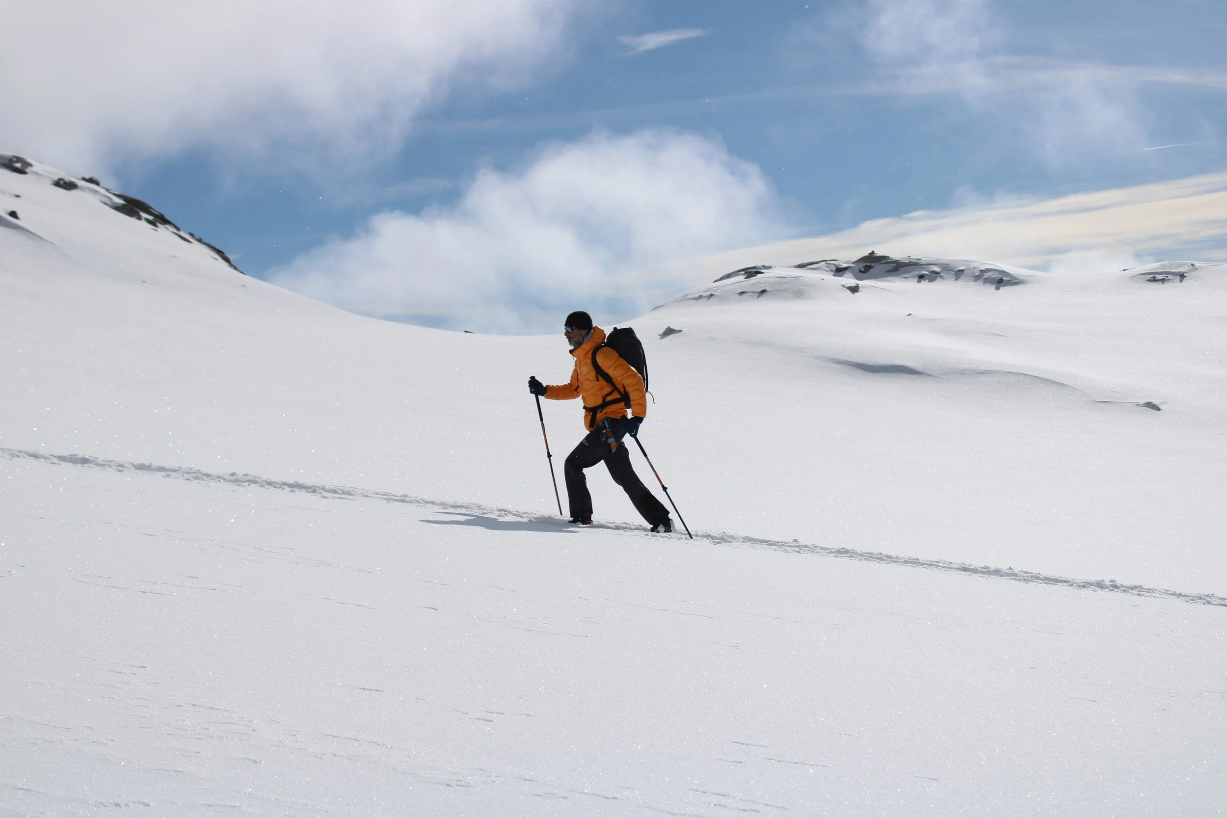 A person wearing an orange jacket, black pants, and sunglasses skiing through snowy terrain under a partly cloudy sky.