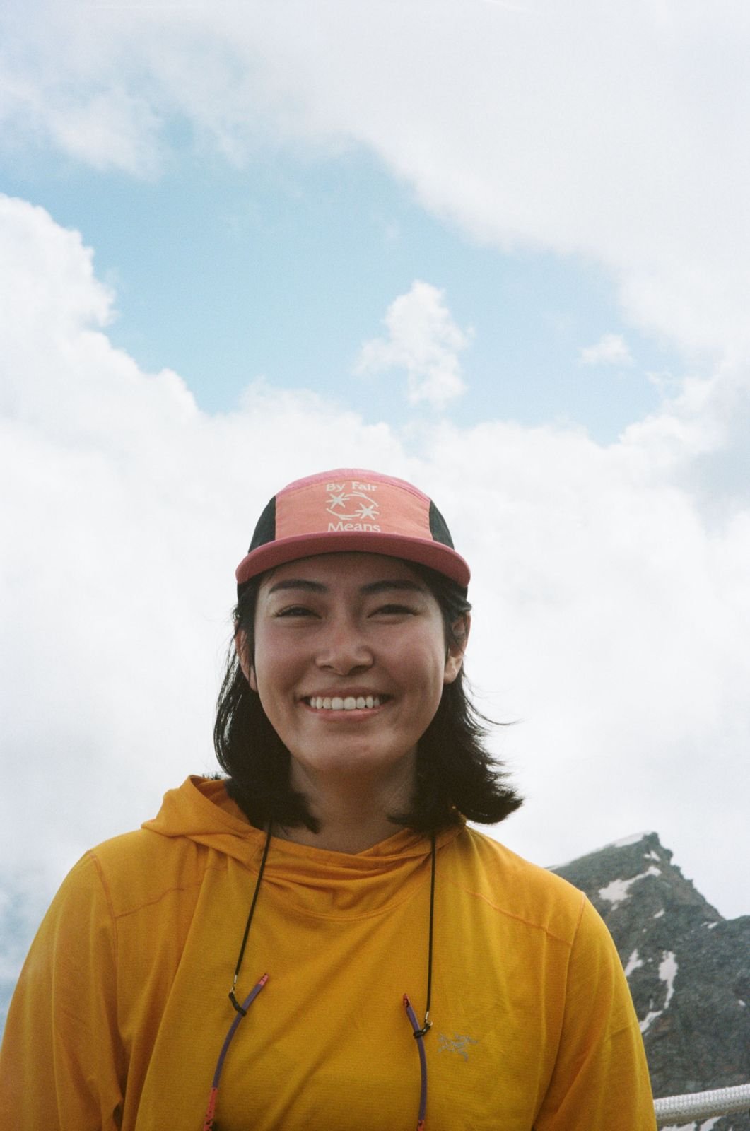 A smiling woman in a yellow jacket and pink cap standing outdoors with cloudy sky and mountain in the background.