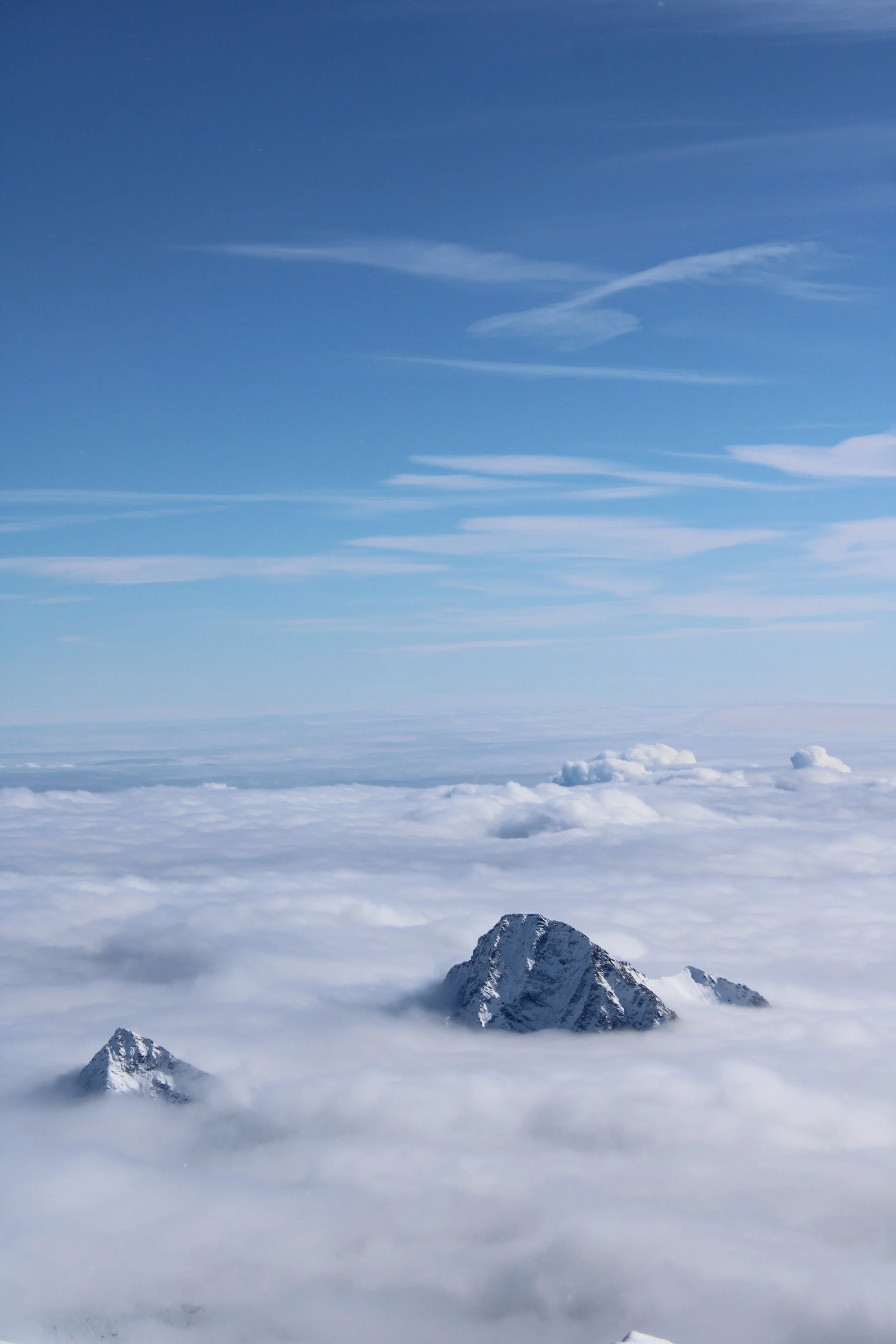 Snow-covered mountain peaks protruding above a thick layer of clouds with a blue sky and wispy clouds overhead.