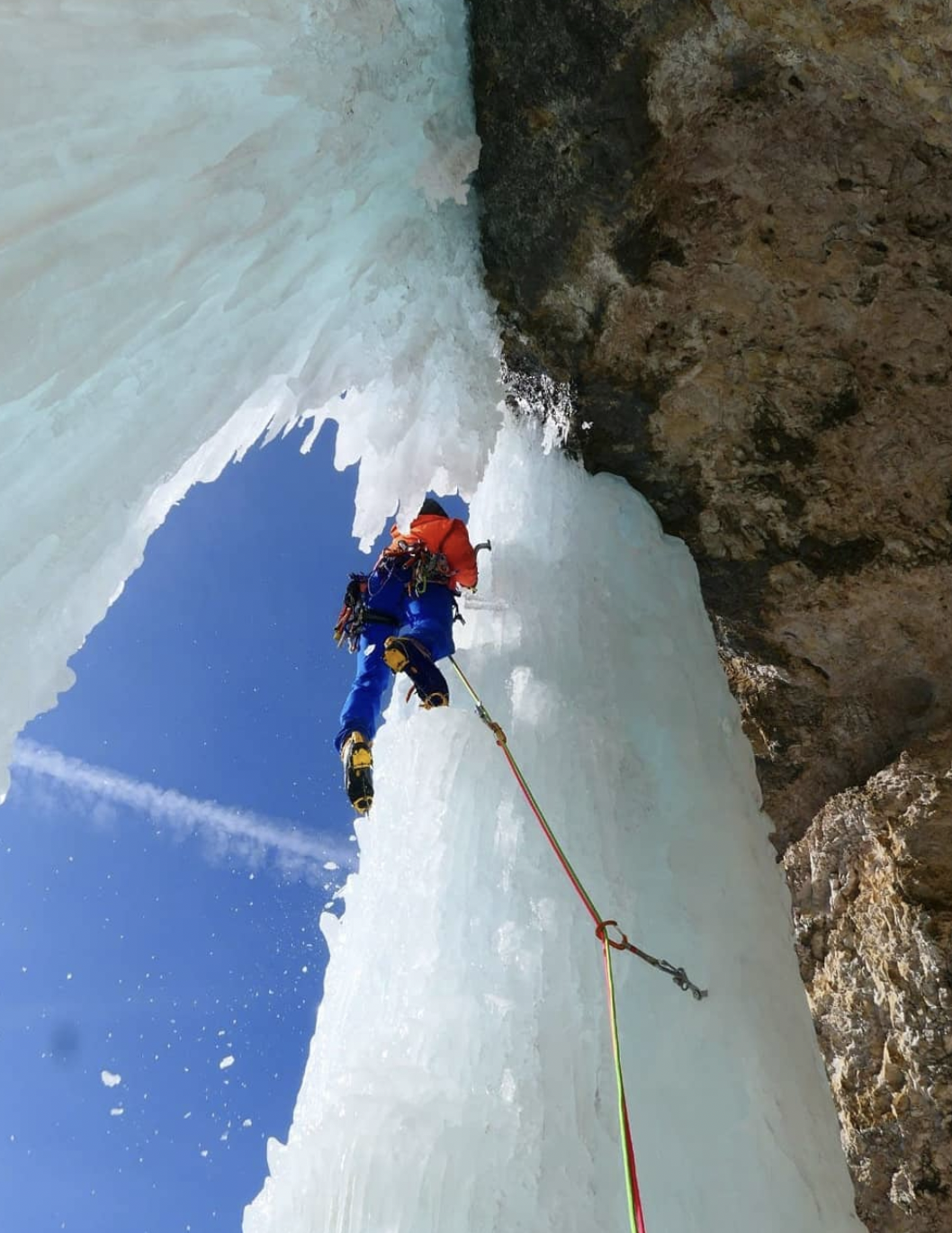 A climber in blue and red climbing gear ascending an ice wall with a clear blue sky in the background.