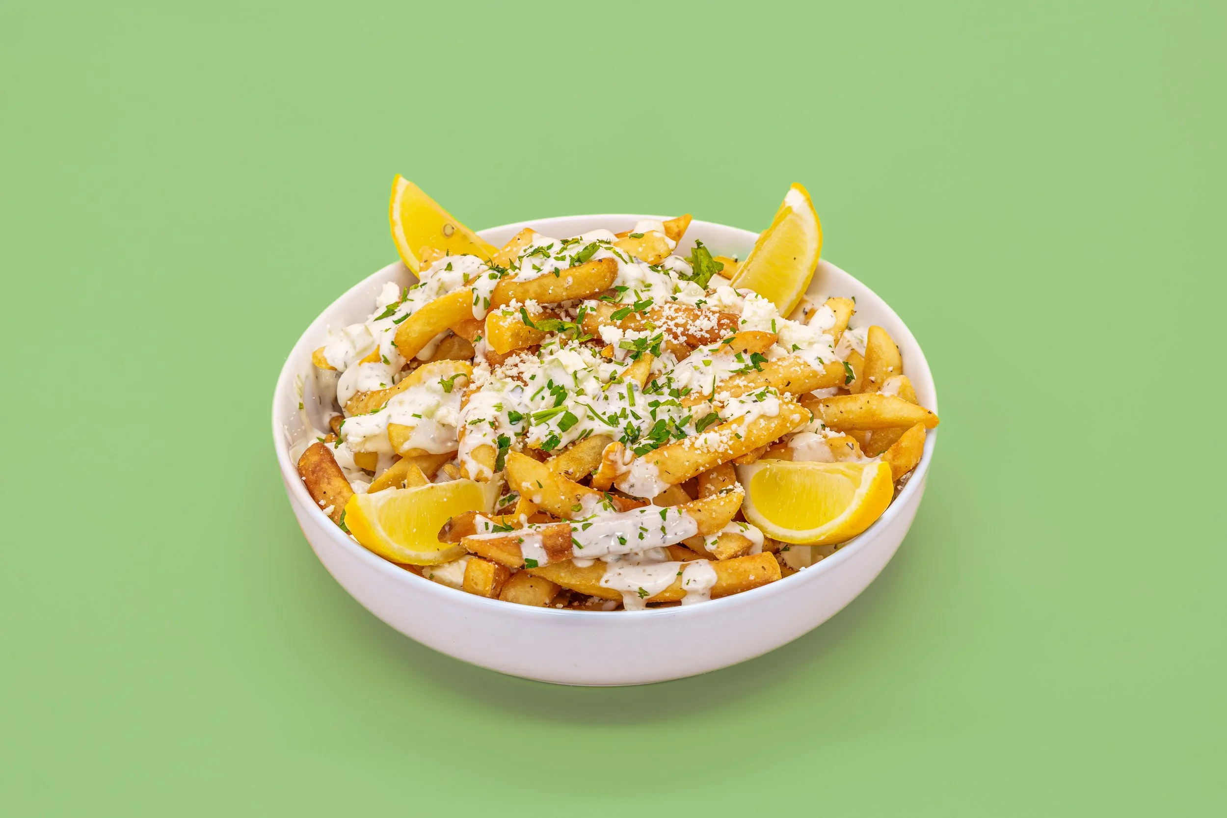 A bowl of poutine topped with gravy, cheese curds, chopped parsley, and lemon wedges on a green background.