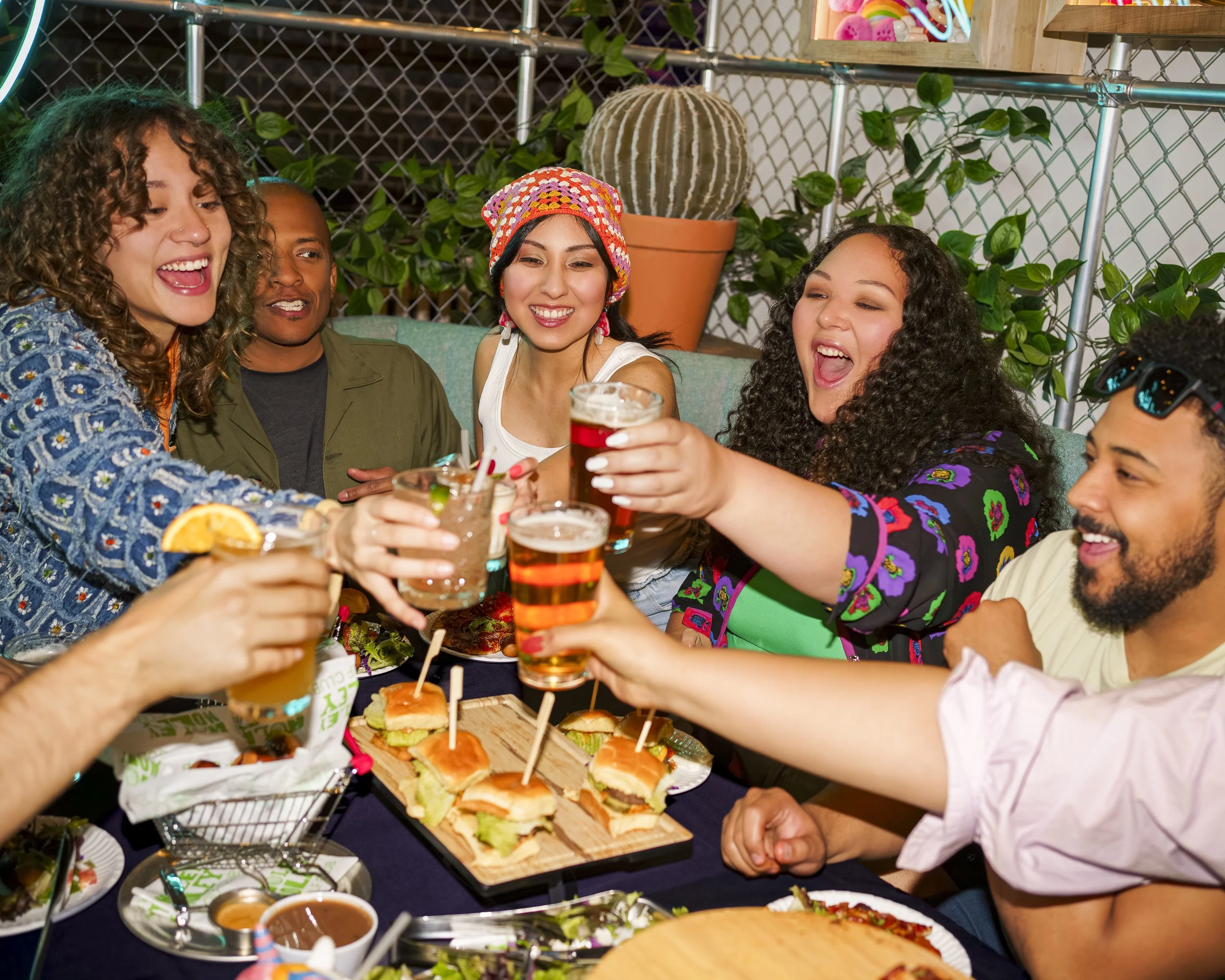 A group of friends celebrating at a party, sitting around a table with sandwiches, drinks, and snacks, raising their glasses and smiling.