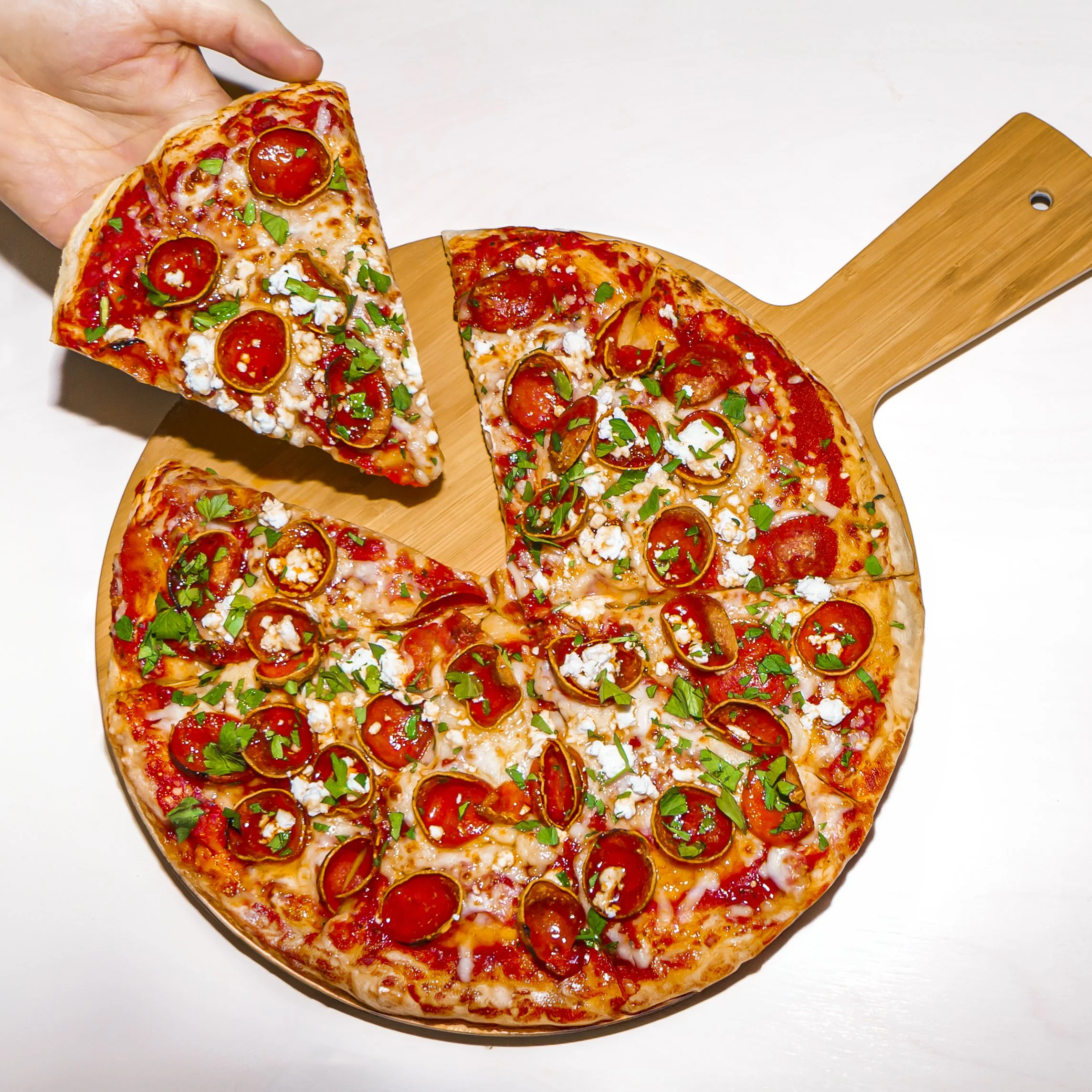 A hand holding a slice of pepperoni pizza with green herbs on top, placed on a round wooden pizza paddle with more slices of pizza in the background.