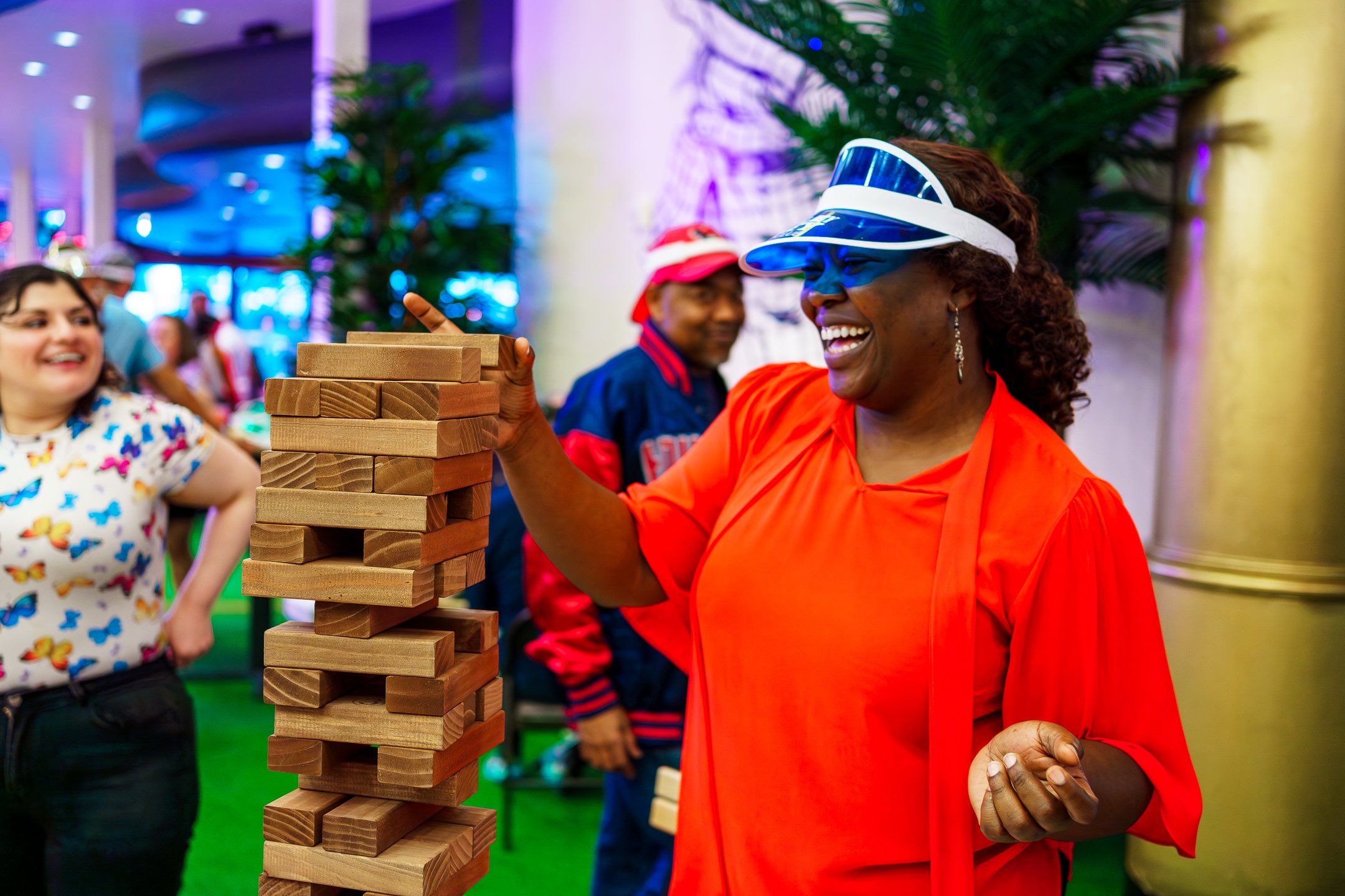 A woman wearing an orange dress and a blue visor hat is playing a giant Jenga game at an indoor event. She is smiling and has dark curly hair, with other people in the background enjoying the activity.