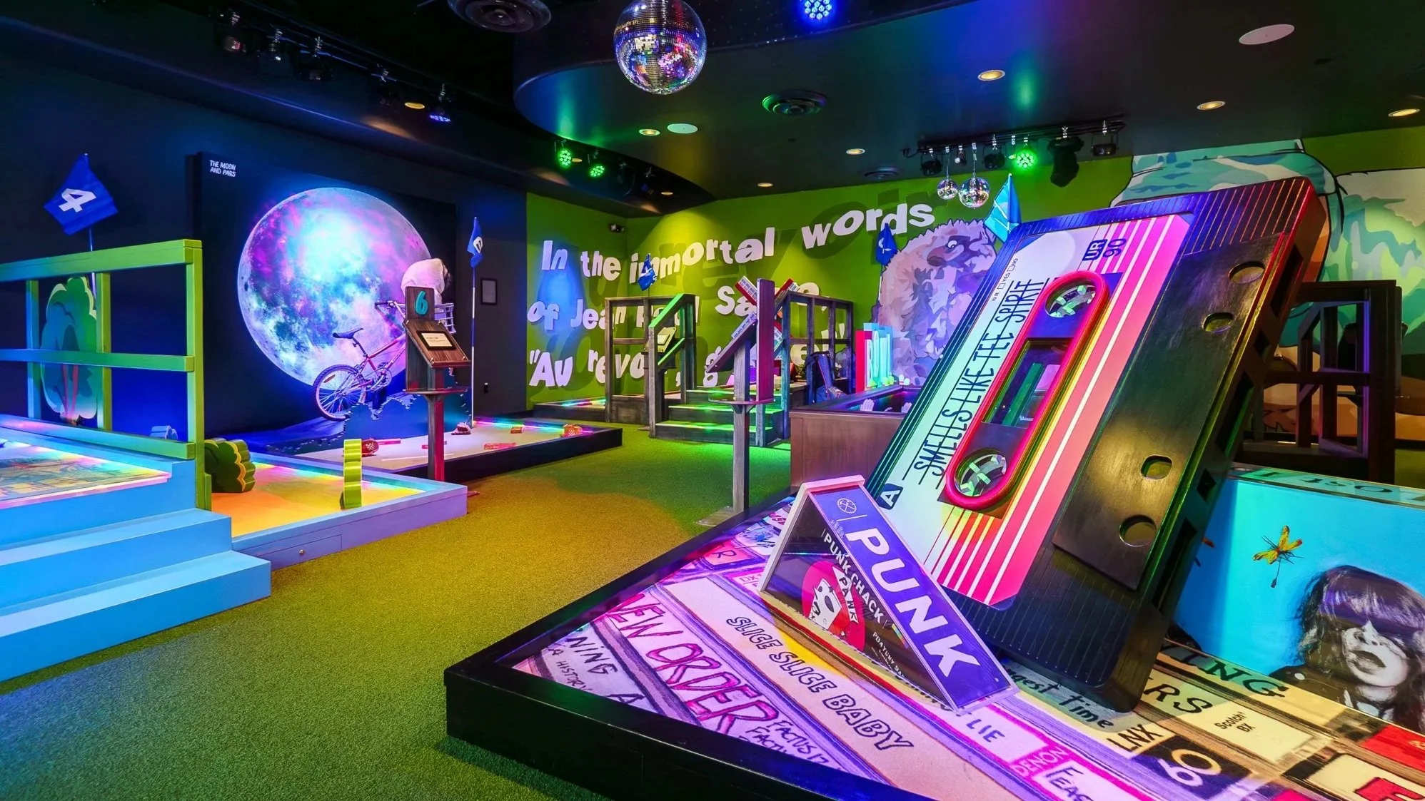 Indoor arcade with colorful neon lighting, paintings of a moon and an eagle on the walls, and various arcade games, including a giant cassette tape game and a giant book with a butterfly decal.