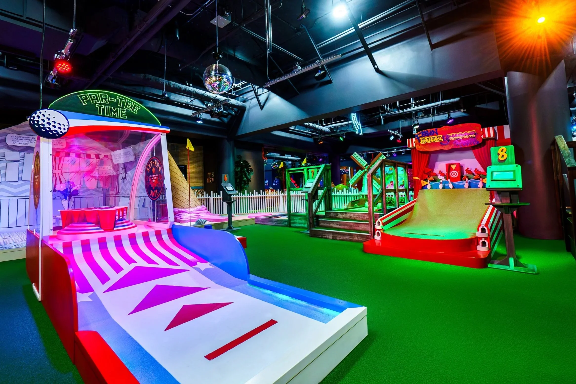 Colorful indoor mini golf course with various game stations, neon signs, bright lighting, and green turf flooring.