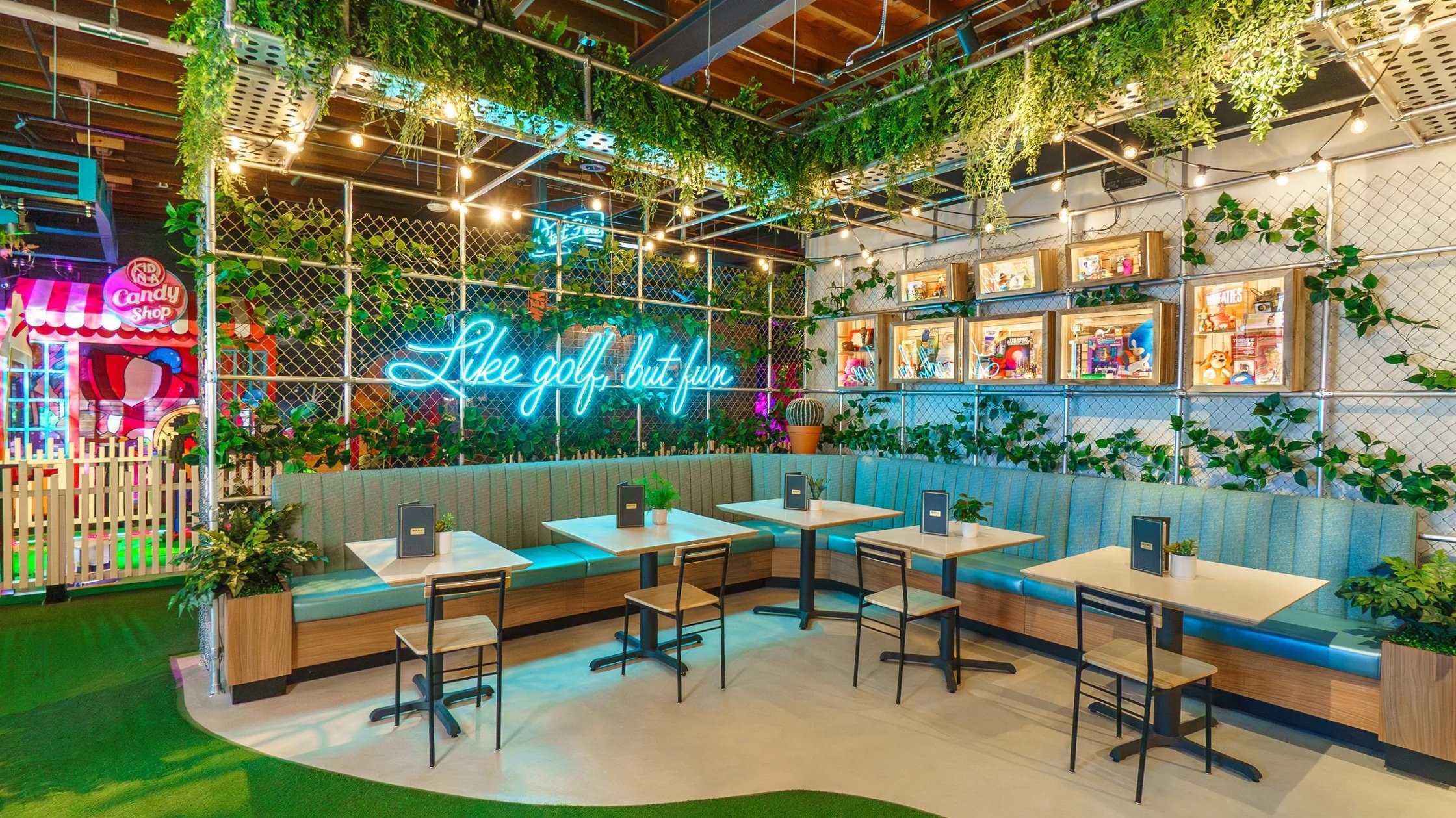 Indoor seating area with light blue upholstered booths, wooden accents, small tables, and black chairs. The space features greenery with hanging plants and vine decorations, a neon sign that reads 'Like golf, but fun,' and framed collectibles and art