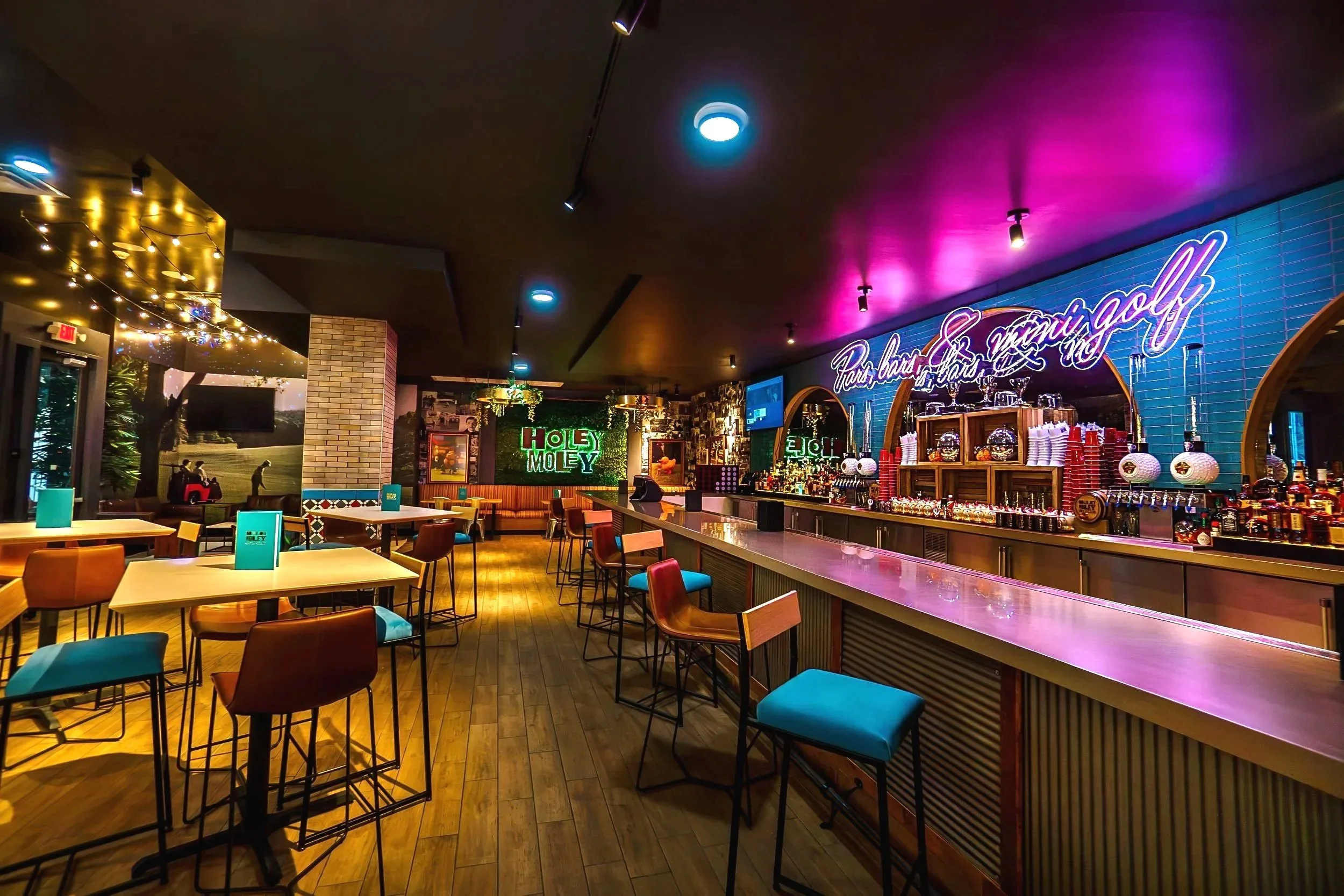 The interior of a bar with colorful lighting, tables and chairs, a long bar counter with drinks, and neon signs.