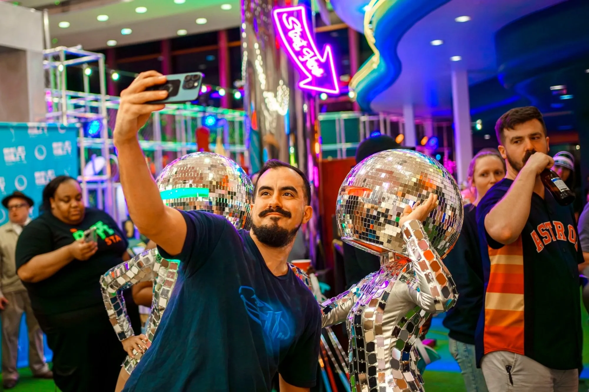People at a vibrant indoor event with colorful neon lights and decorations. A man is taking a selfie, and two others are holding mirror-covered sculptures. The background includes a neon sign and several attendees.