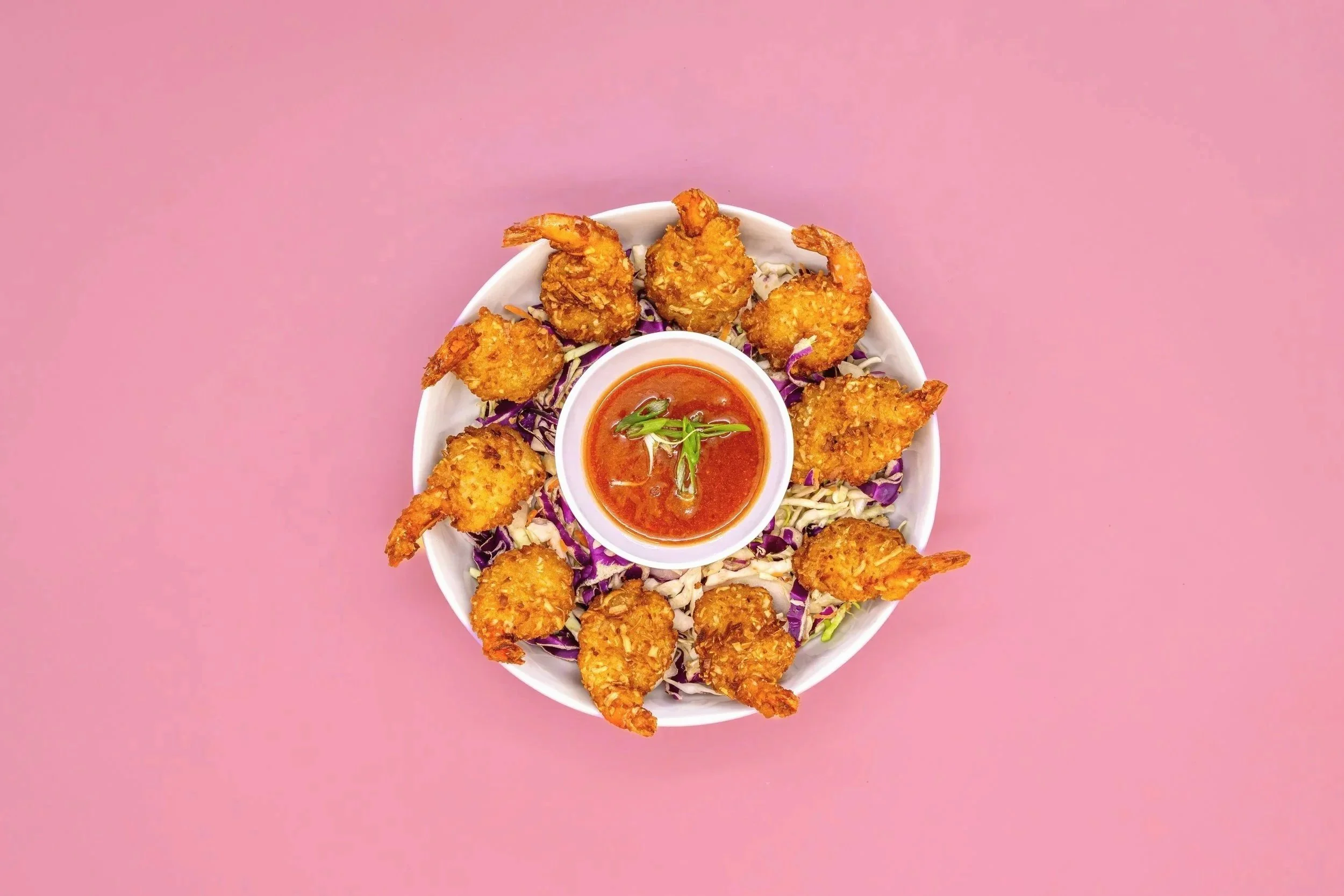 A white bowl of fried shrimp with breaded coating, served with a small cup of red dipping sauce garnished with green onion, on a bed of shredded purple and white cabbage, against a pink background.