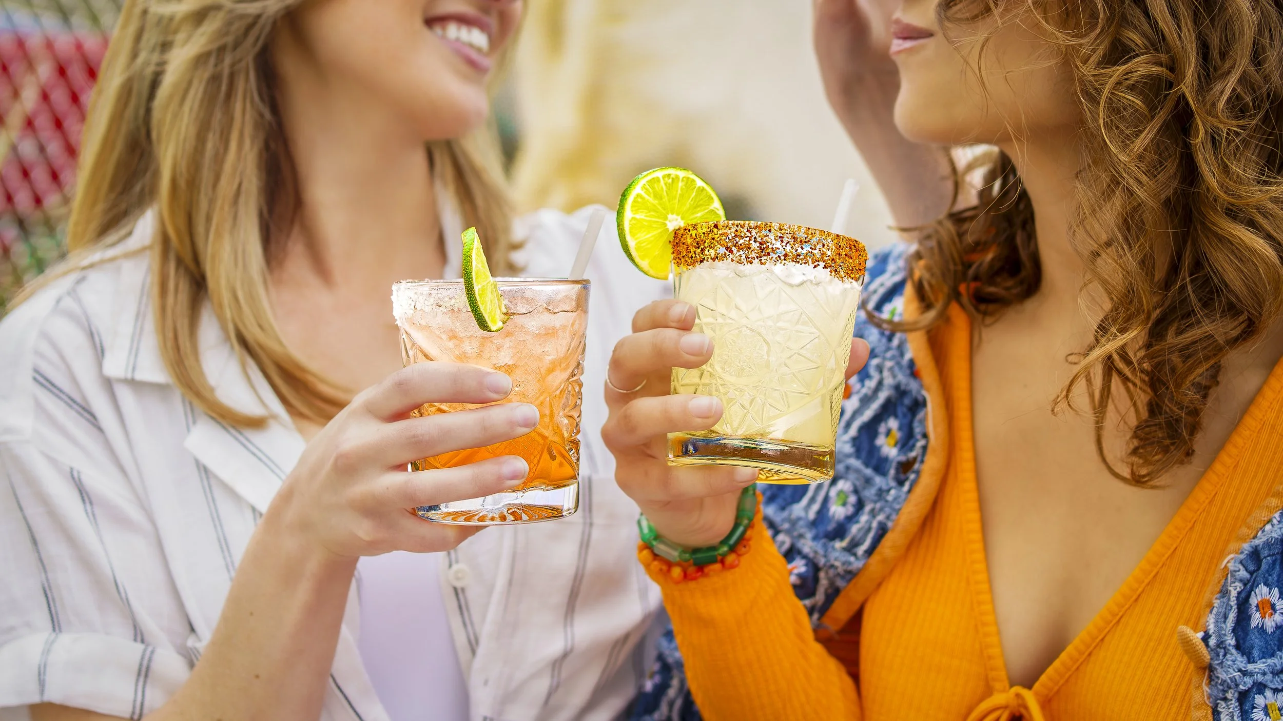 Two women clinking glasses with colorful cocktails topped with lime slices, one with chili powder rim and the other with sugar rim, smiling at each other.