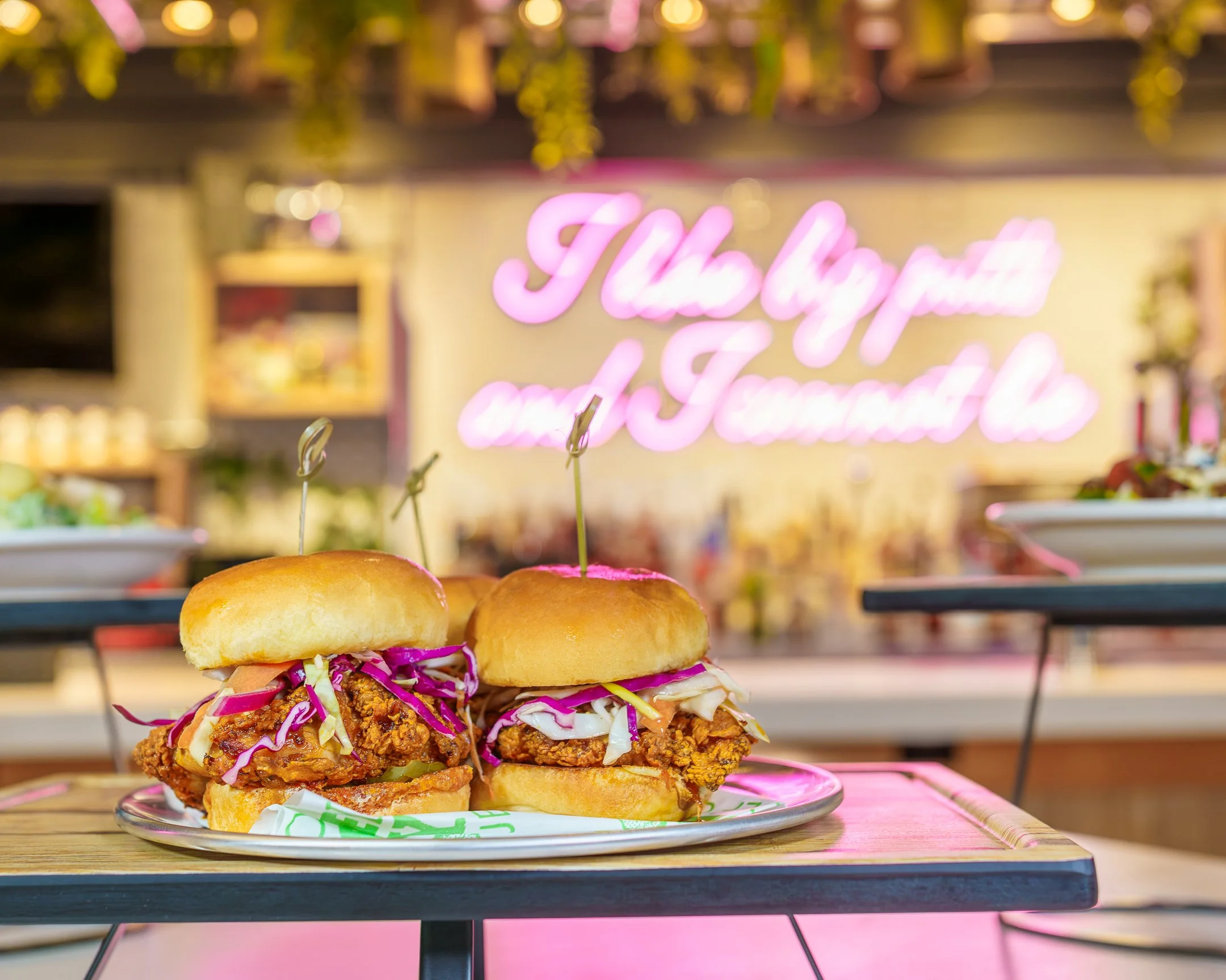 Two fried chicken sandwiches with coleslaw and pickles on a plate, set on a wooden table at a restaurant with a blurred background featuring a pink neon sign and hanging plants.