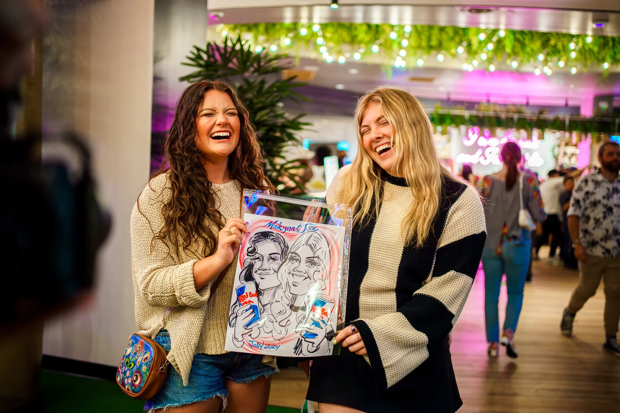 Two young women stand together in a lively indoor space, laughing and smiling as they hold a colorful caricature artwork of themselves, with a crowded background decorated with green plants, hanging string lights, and pink neon lighting.