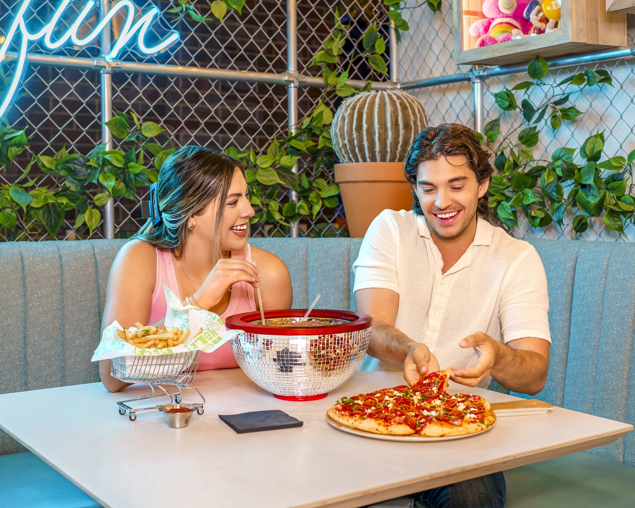 A young man and woman sitting at a table in a pizzeria, enjoying slices of pizza, with a pizza box and a bucket of fries on the table. They are smiling and appear to be having a good time. There are green plants and decorative elements on the wall be