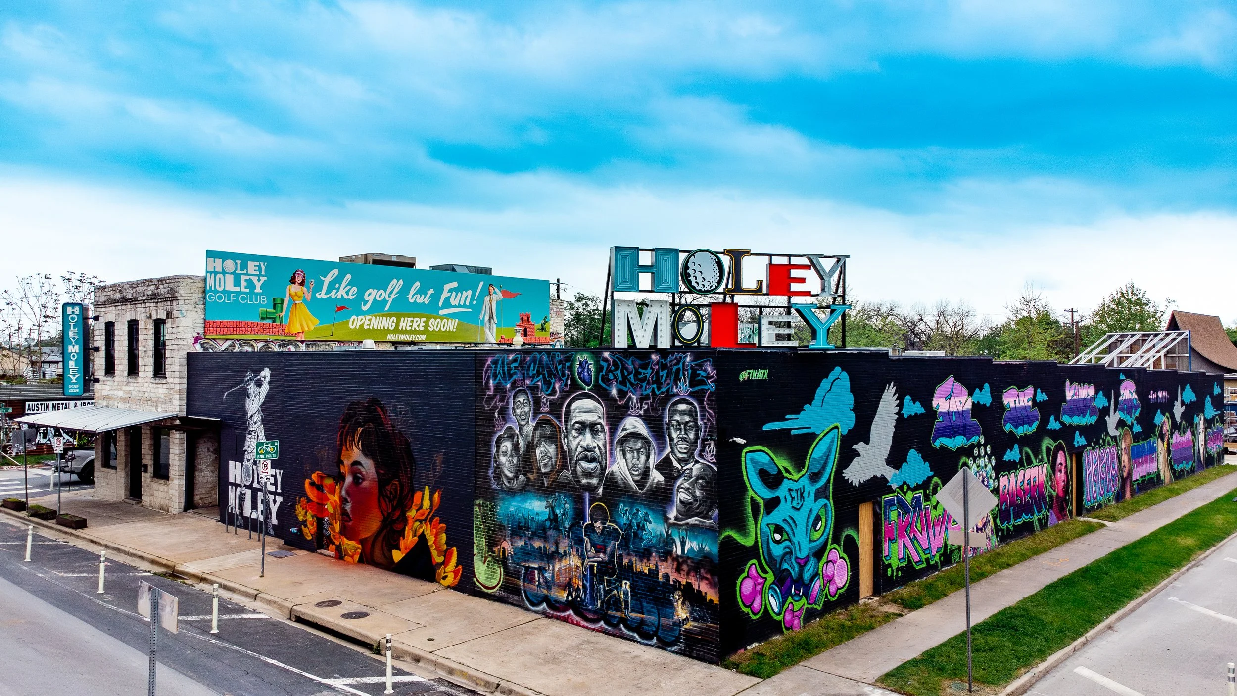 A building decorated with colorful graffiti art and murals, features a large sign that says 'Holey Moley' and a billboard promoting a golf course with the slogan 'Like golf but Fun!' The area is outdoors with a sidewalk, some trees, and a street with white posts.