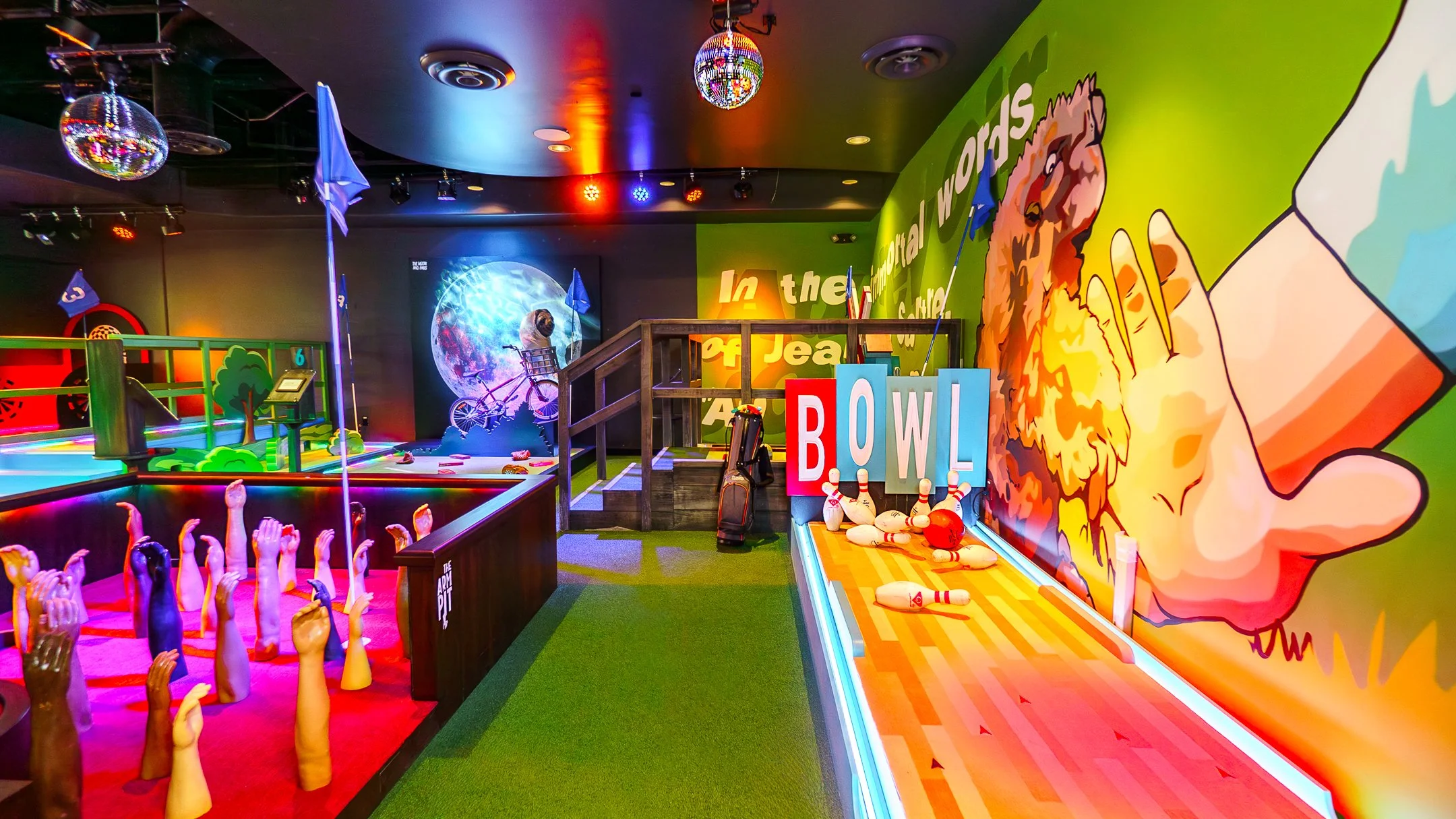 An indoor arcade with colorful neon lights and vibrant wall murals. There is a bowling area with pins knocked down, a skee-ball game with a large mascot, and a safety signage. The ceiling has disco balls and colored lights, creating a lively atmosphe