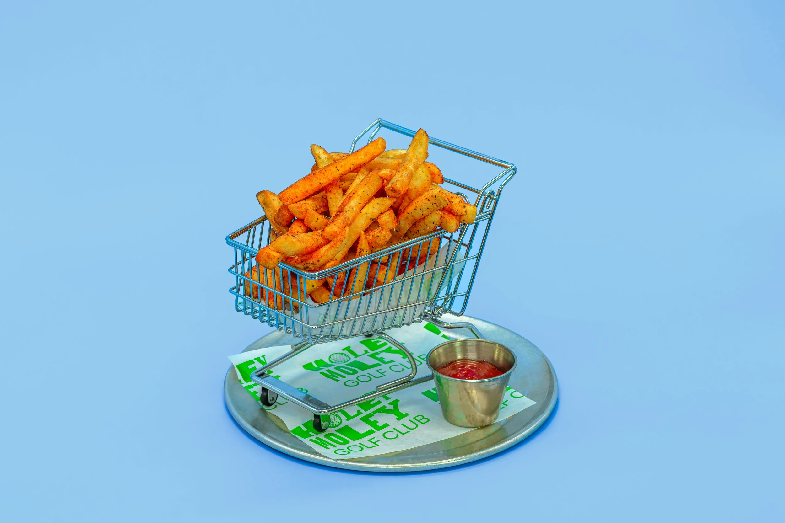 French fries in a small metal shopping cart on a round tray with a cup of ketchup, with golf course advertising paper underneath, against a bright blue background.