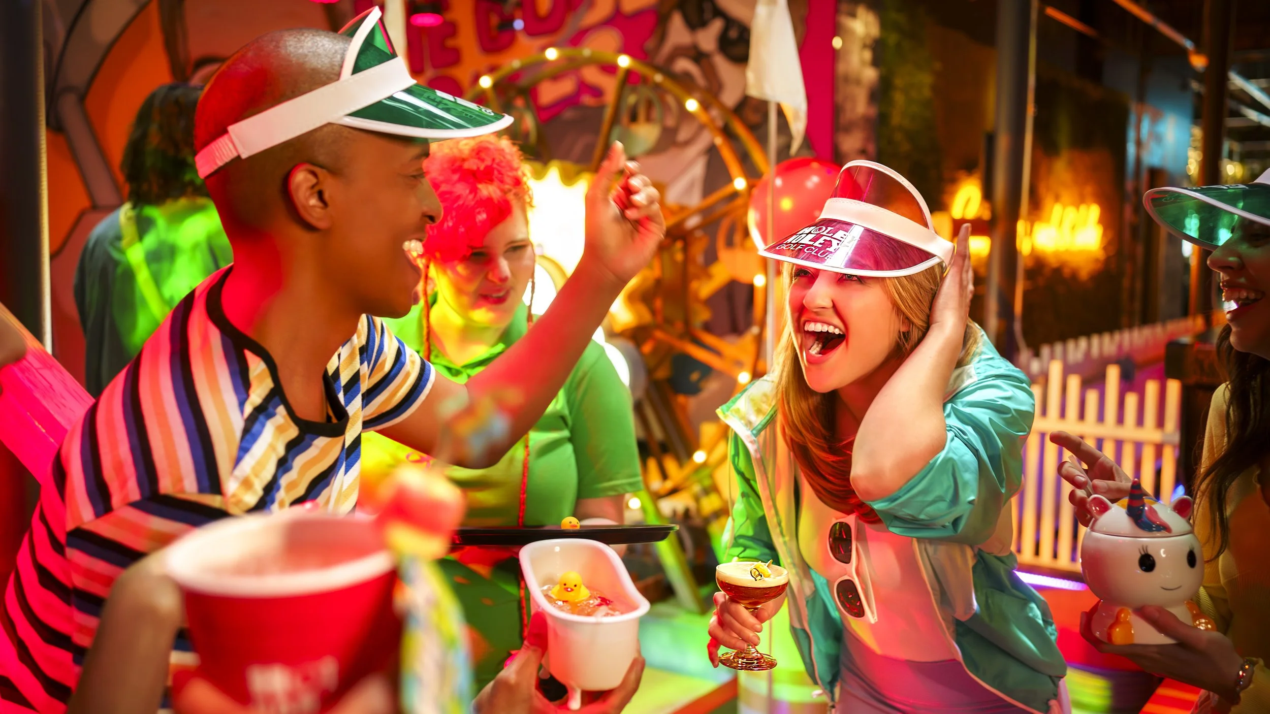 Group of young people celebrating at a colorful carnival or fair, wearing neon and striped clothing, with carnival games and bright lights in the background.