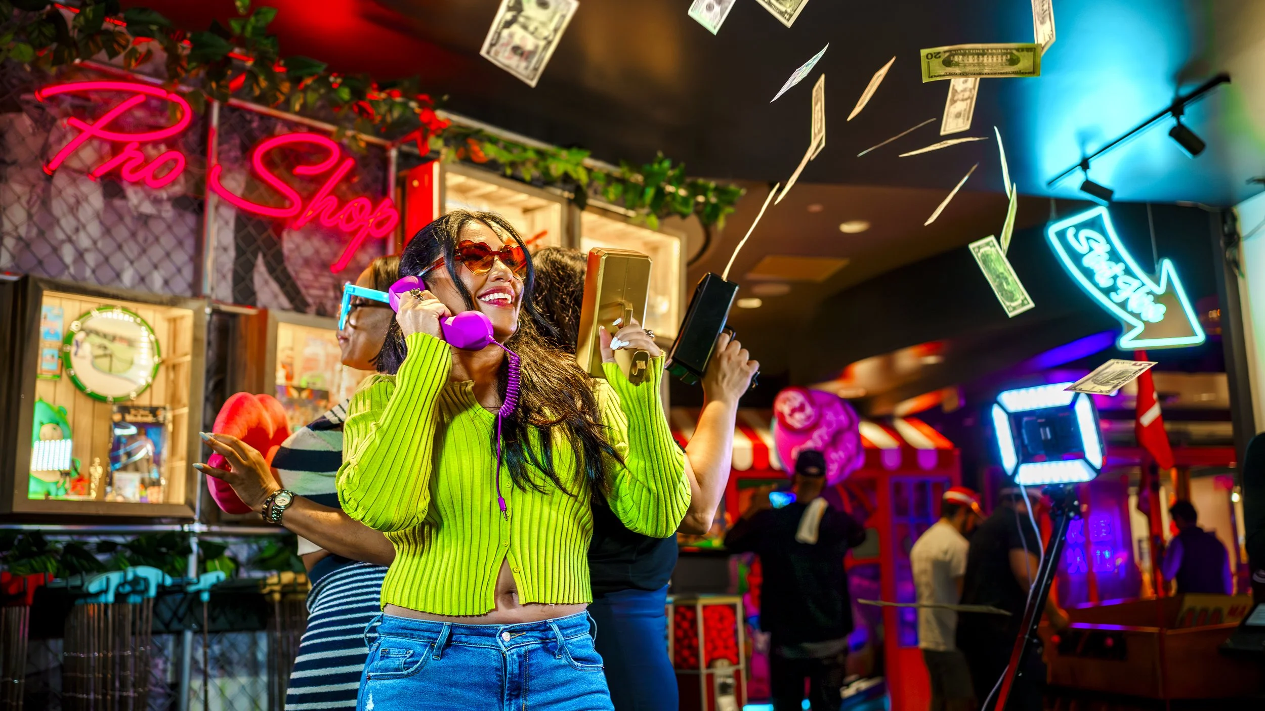 A woman smiling and talking on her pink rotary phone at an arcade or gaming center, with fake money falling around her, colorful neon signs in the background, and other people visible in the setting.