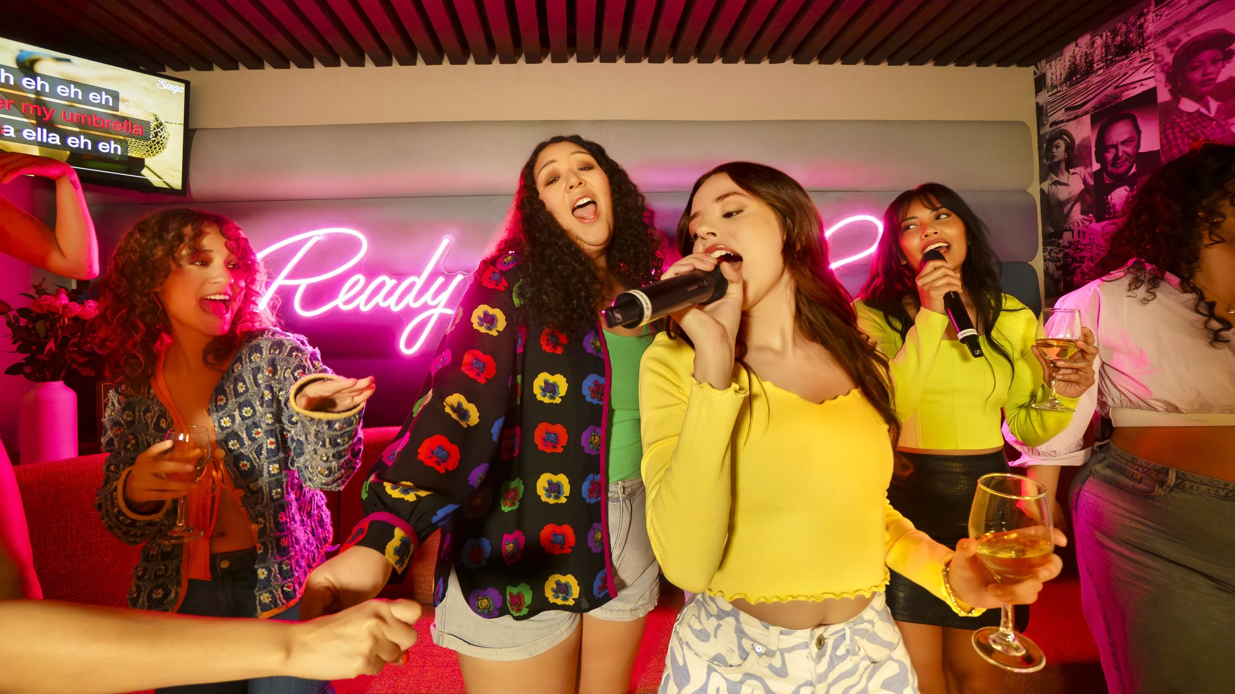 A group of women singing karaoke and holding drinks in a lively, colorful room with neon lights and photo murals.