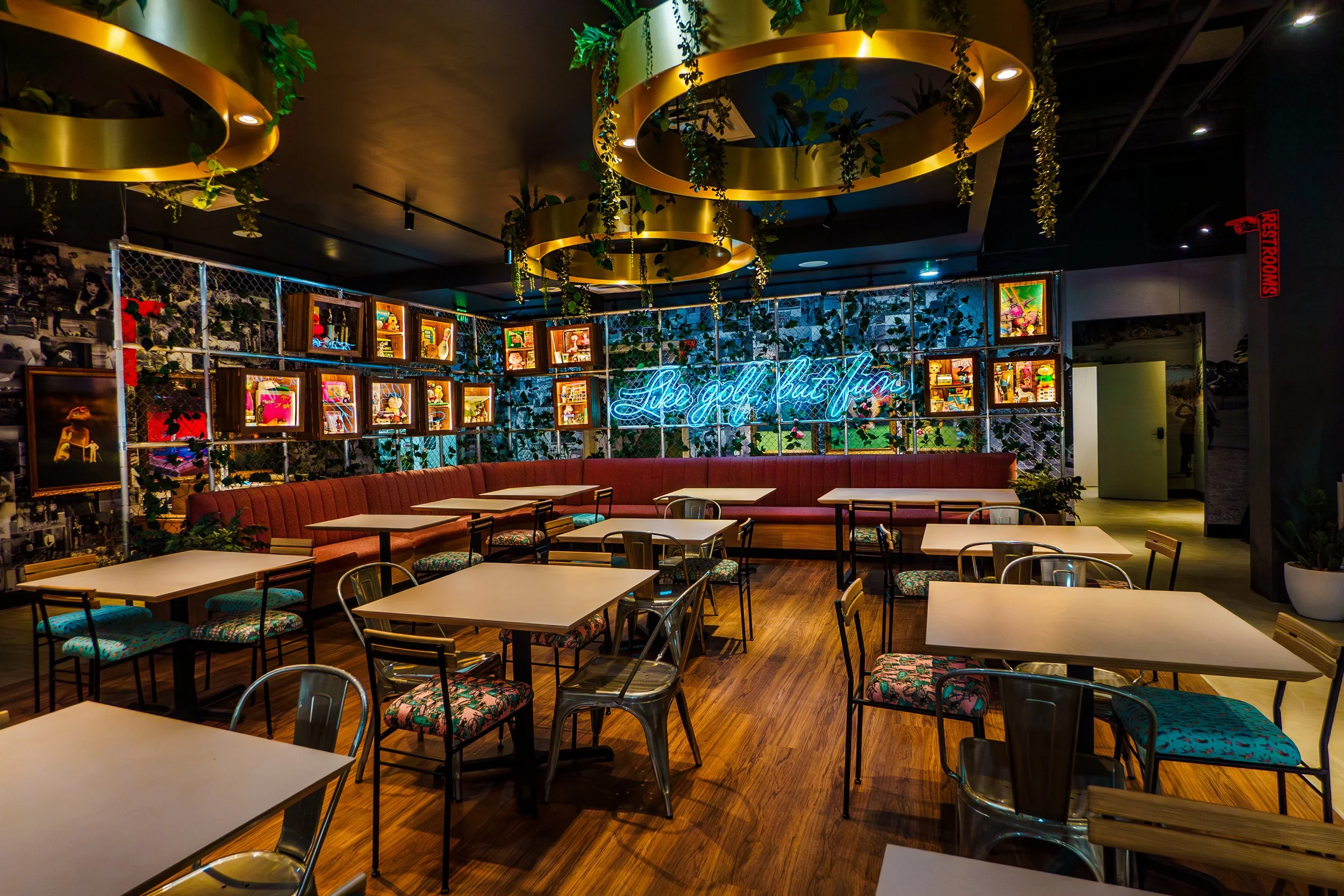Interior of a modern restaurant or bar with white tables, colorful chairs, decorative greenery, and gold circular ceiling fixtures, with a neon sign that reads 'like golf but fun' in blue.
