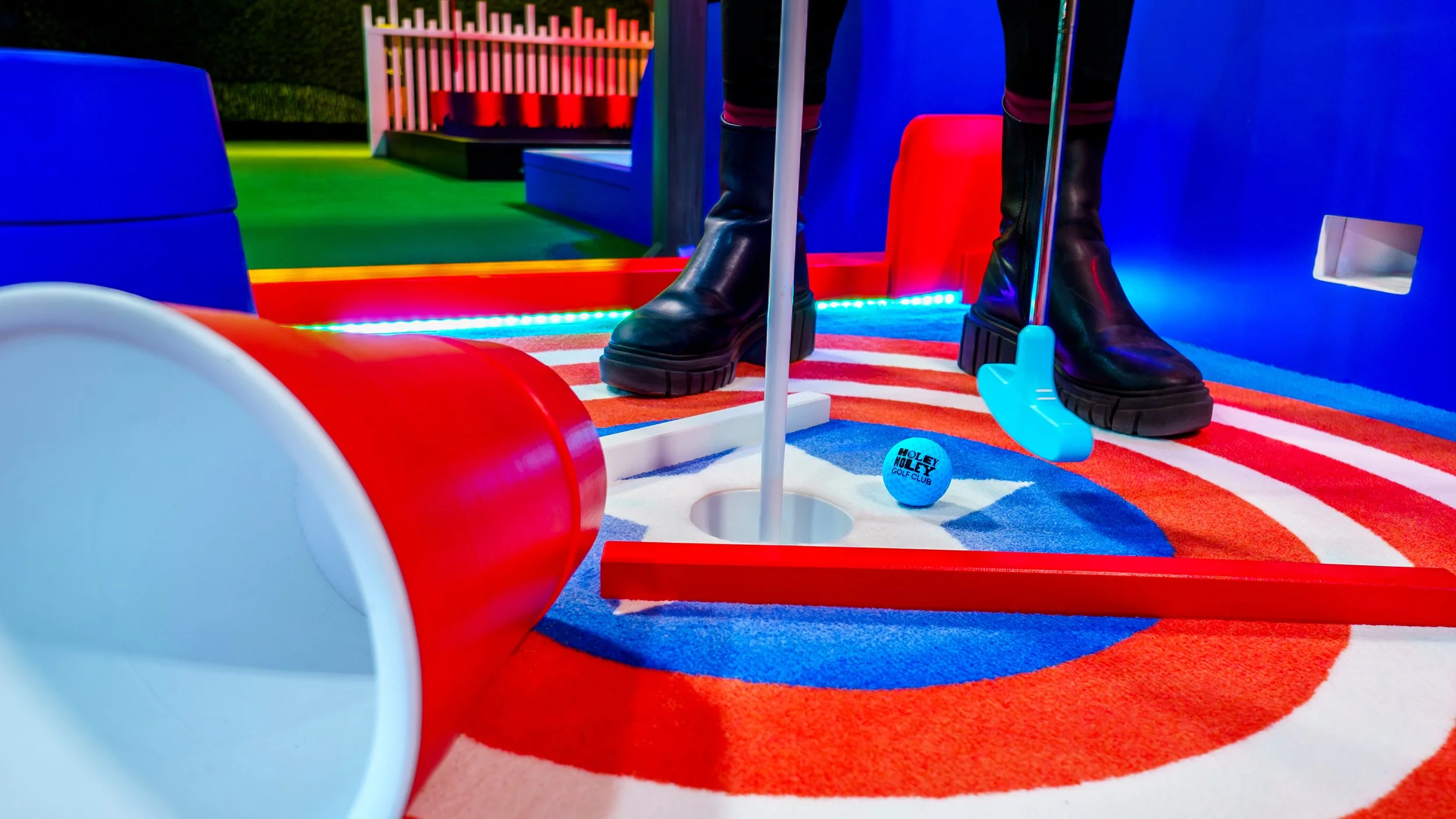 Close-up of a mini golf setup with a blue ball labeled 'Holey Moley Golf Club' near a hole, a red obstacle, and a putter. Colorful lighting and striped rug in the background.