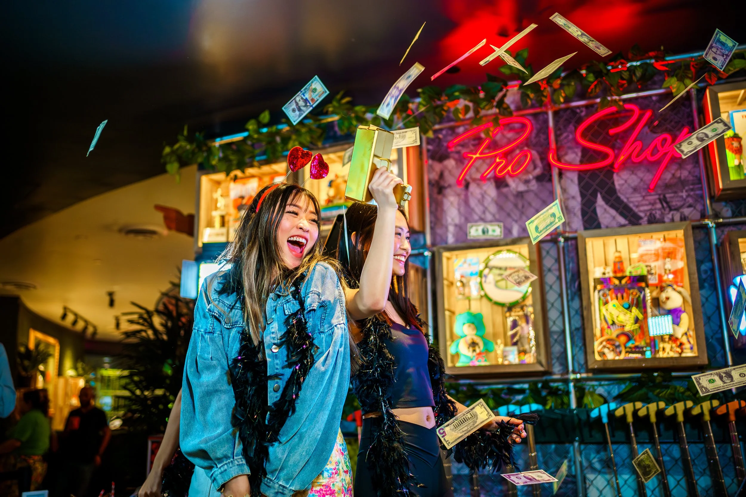 Two women are celebrating at an arcade or carnival with money falling around them. One woman is smiling broadly and holding up a gold-colored toy gun or cash cannon. The background features bright neon lights and the sign reads 'Pro Shop'.