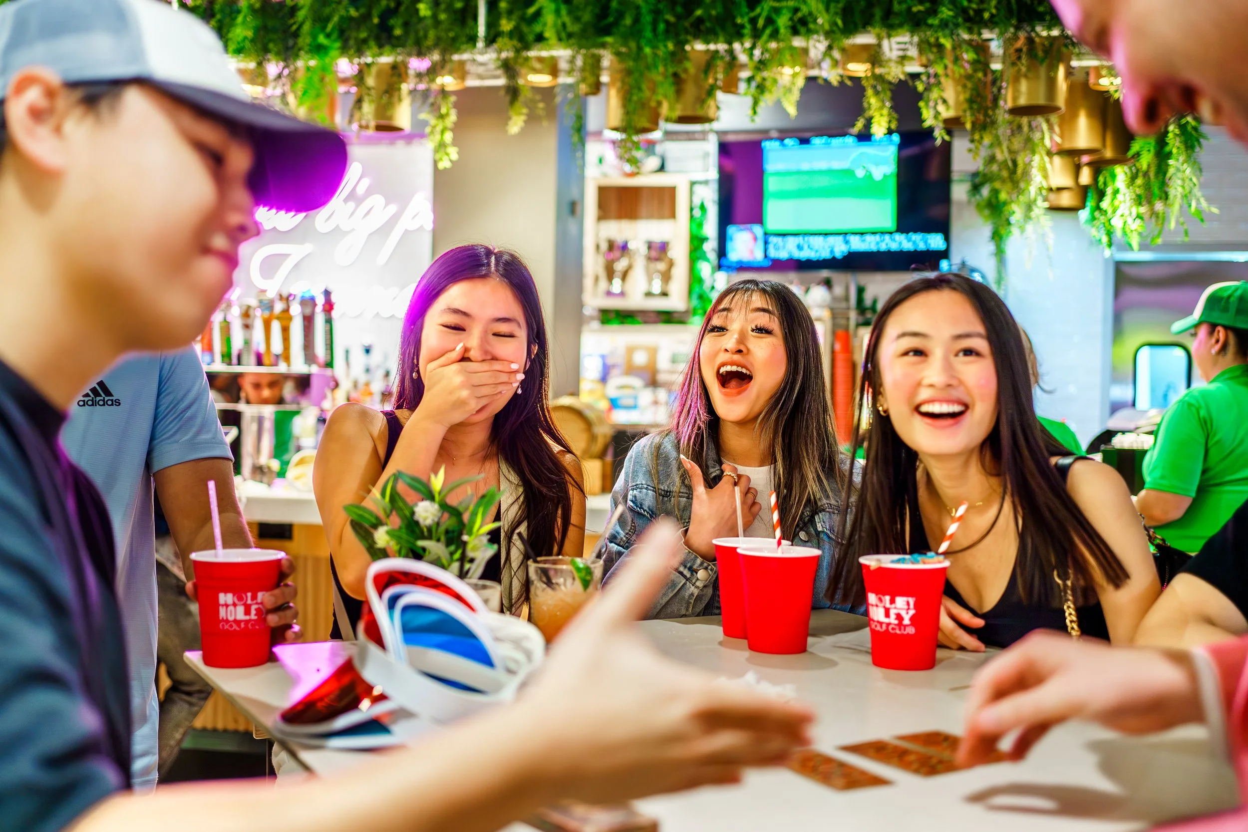 Group of friends laughing and enjoying drinks at a coffee shop or restaurant with green hanging plants and digital screens in the background.