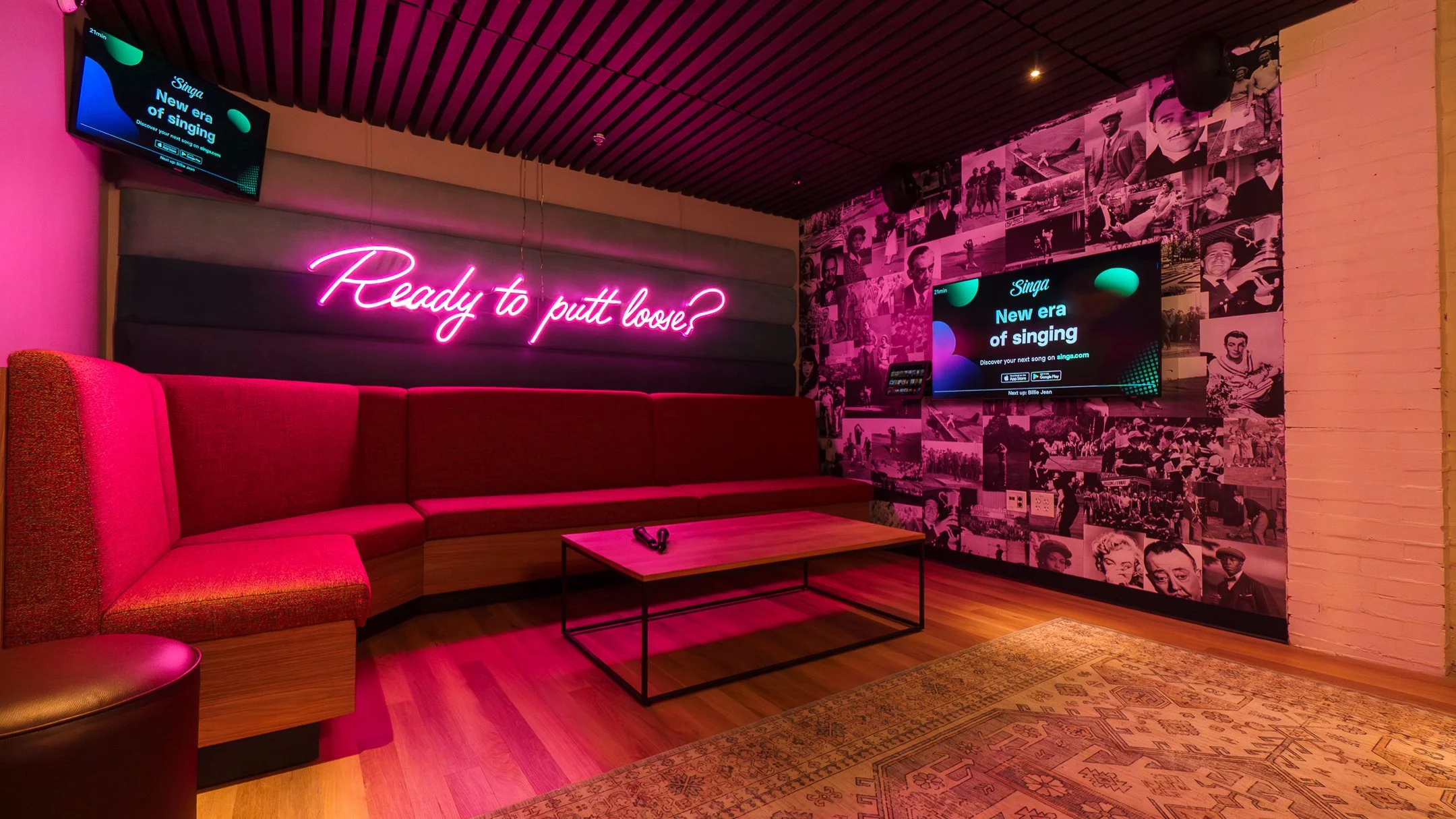 A cozy lounge with a pink neon sign that says "Ready to putt loose?" on a padded wall, a red sectional couch, a wooden coffee table, and two large digital screens displaying an advertisement for Singa's new era of singing, surrounded by a collage of 