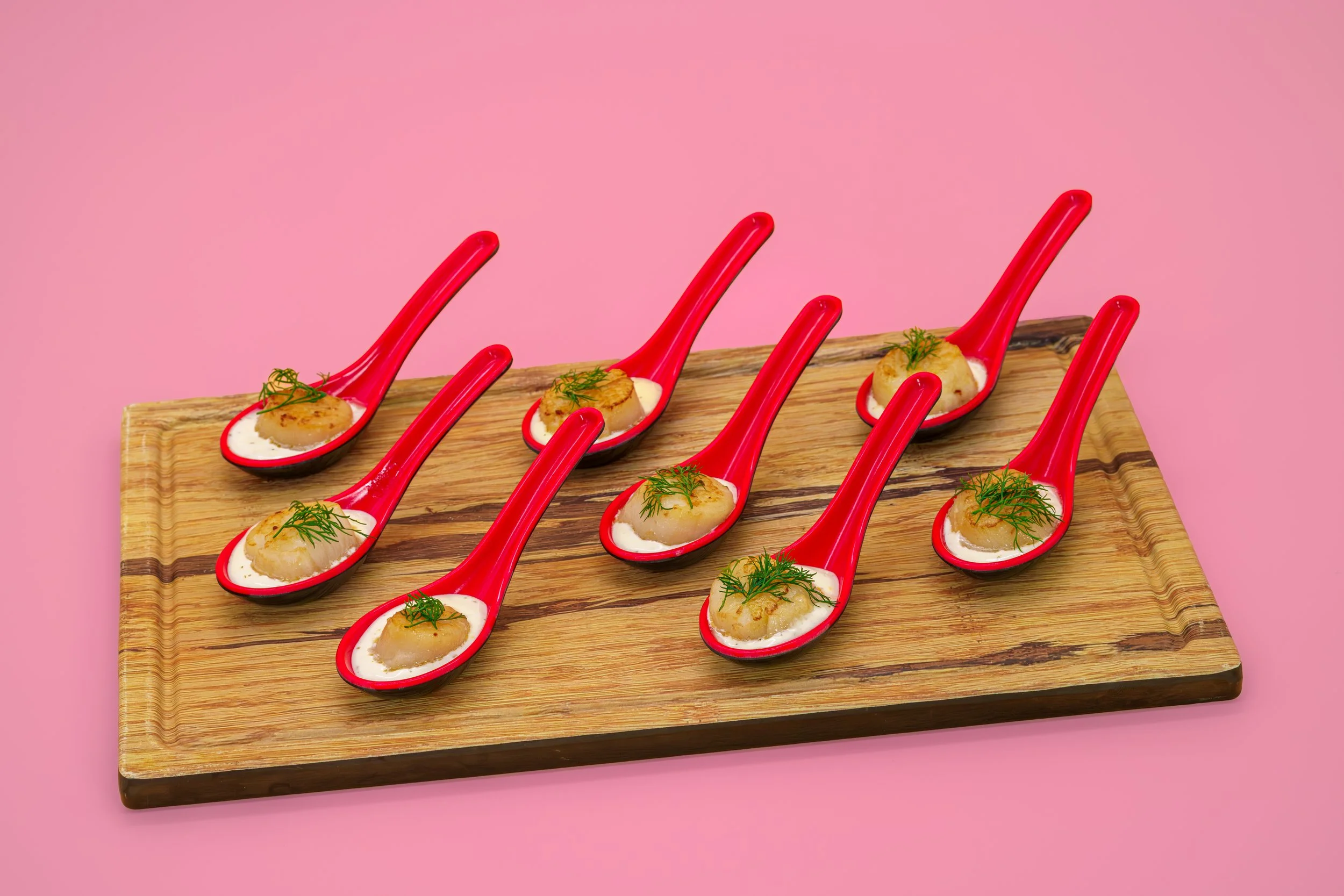Ten red ceramic spoons filled with scallops, topped with herbs, arranged on a wooden serving board against a pink background.