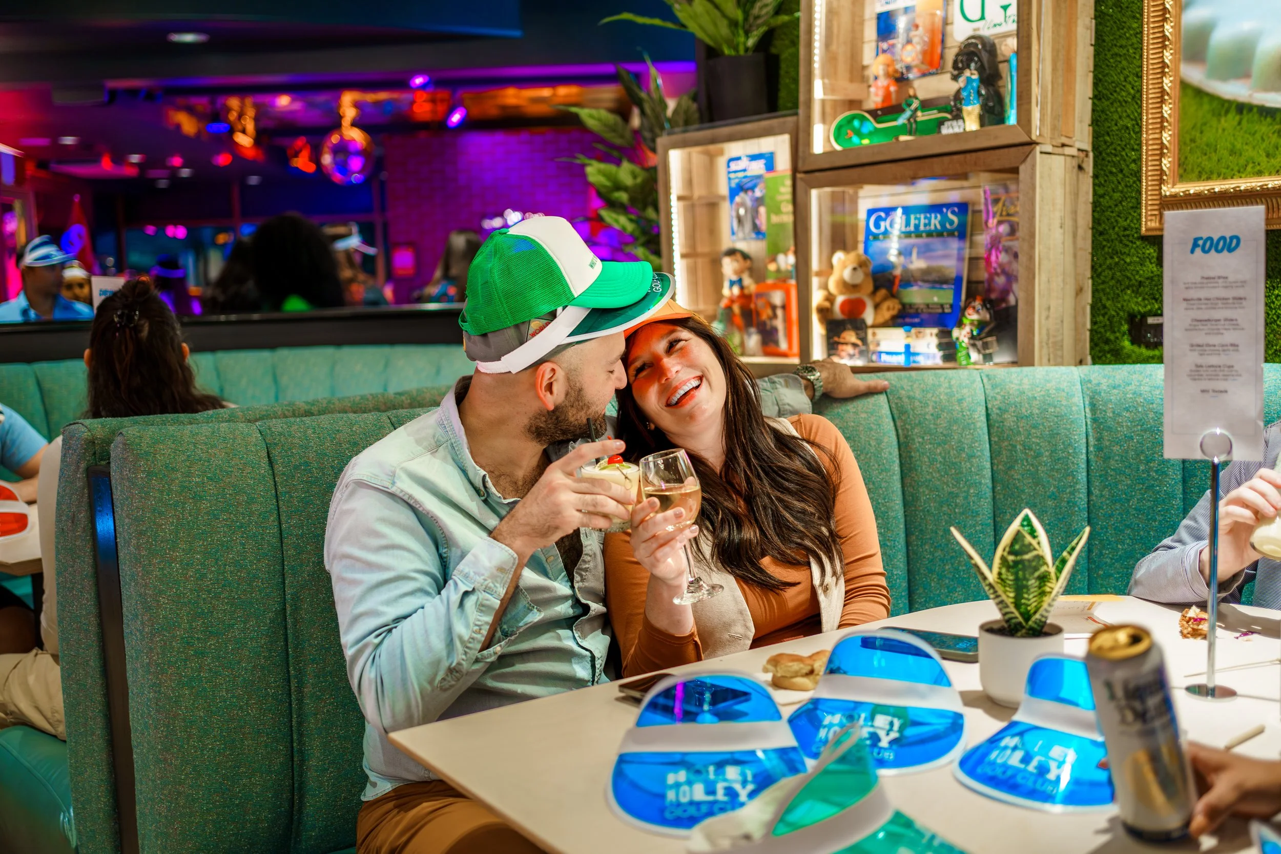 People dining in a lively, colorful restaurant with neon lighting, drinks, and themed decor, including framed photos, toys, and a sign reading 'FOOD'.