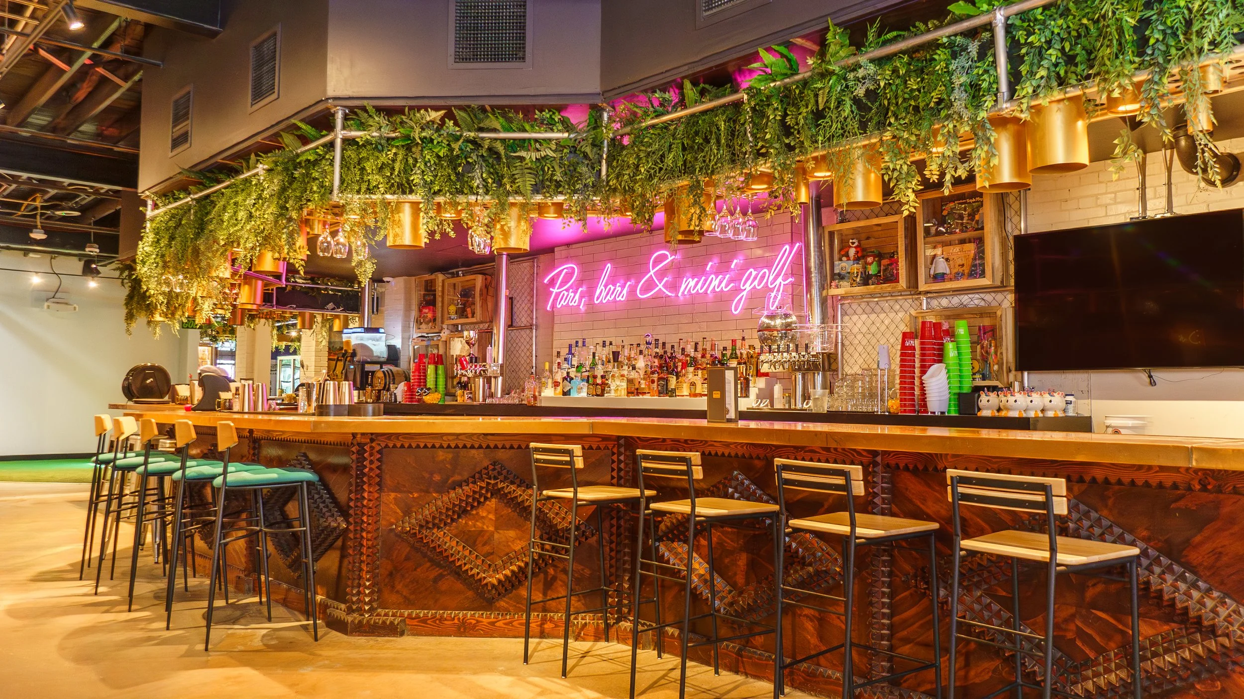 Bar with a neon pink sign that says 'Paris, bars & mini golf', decorated with hanging green plants and gold light fixtures, featuring bar stools, glasses, bottles, and a TV screen.