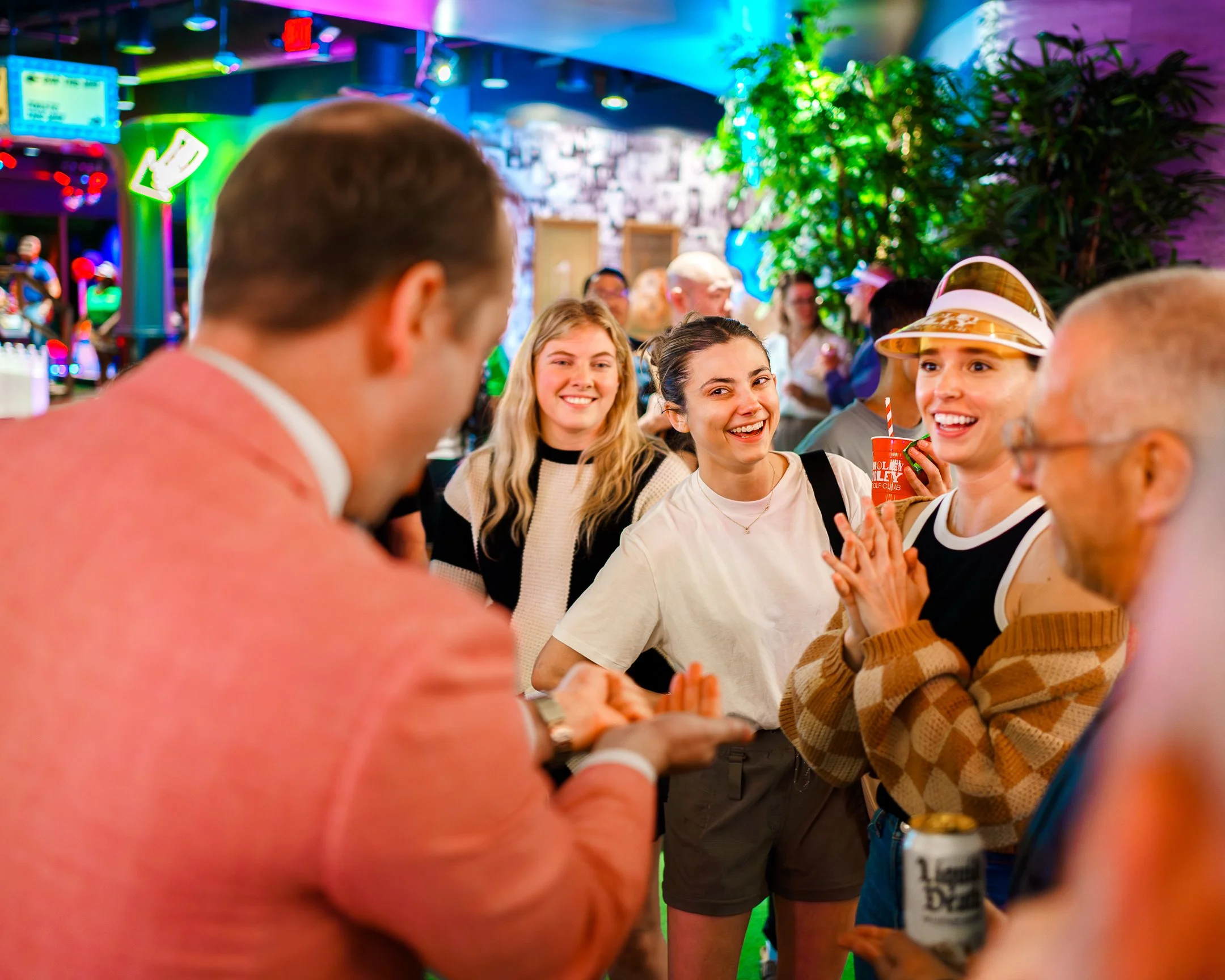 People socializing at a lively indoor party with colorful neon lights, a large green plant, and a wall decorated with black-and-white photos.