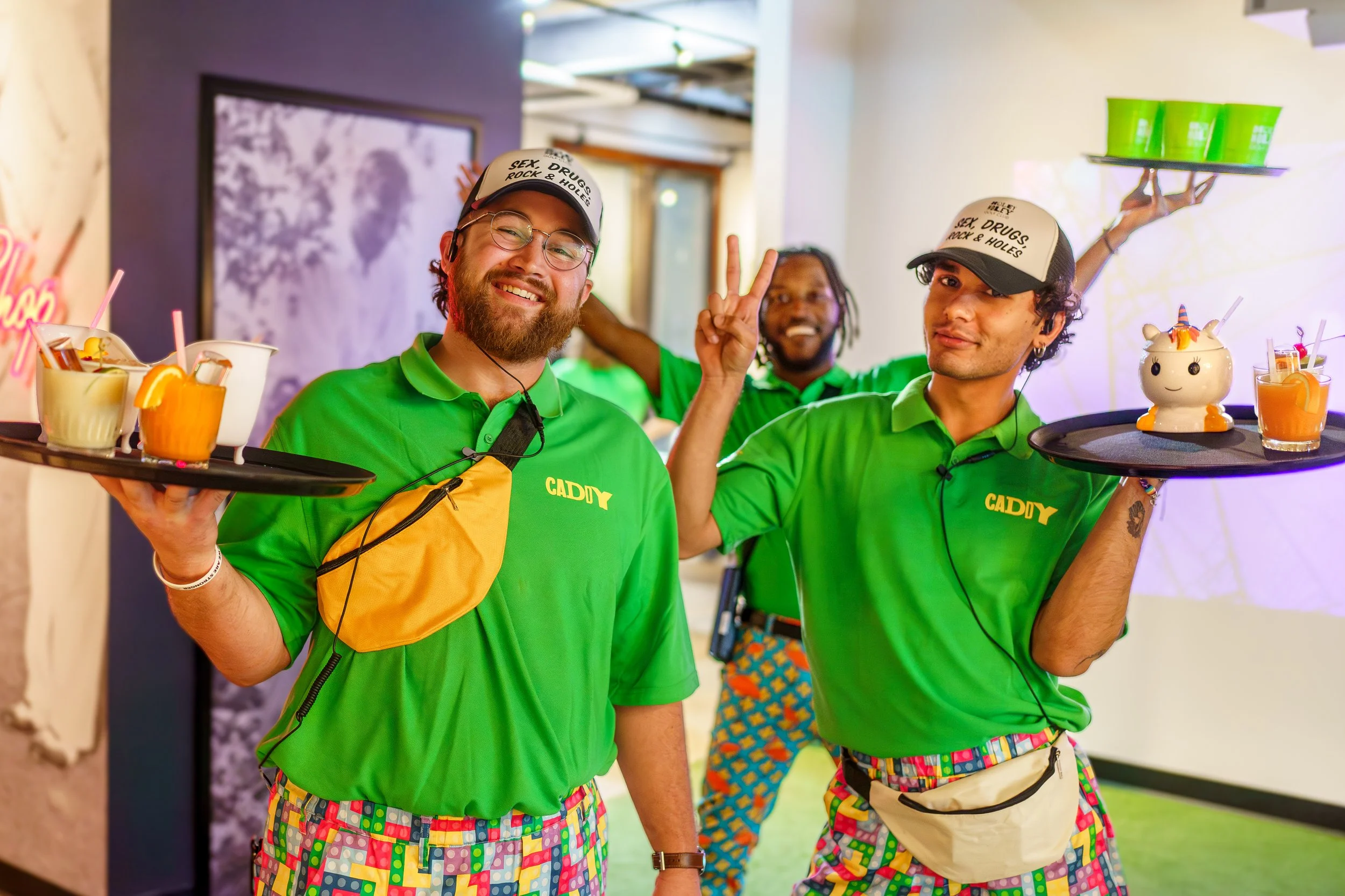 Three smiling men in green shirts with colorful patterned shorts and hats, holding drinks and trays, inside a lively decorated venue.
