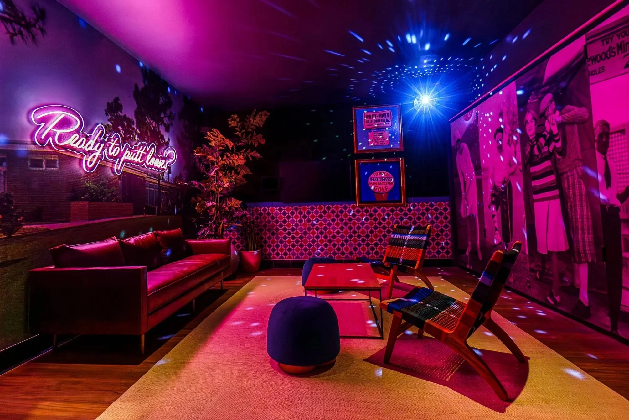 A cozy indoor lounge area with purple and pink neon lighting, a red leather sofa, modern chairs, a yellow rug, and wall art of mannequins and vintage advertisements.