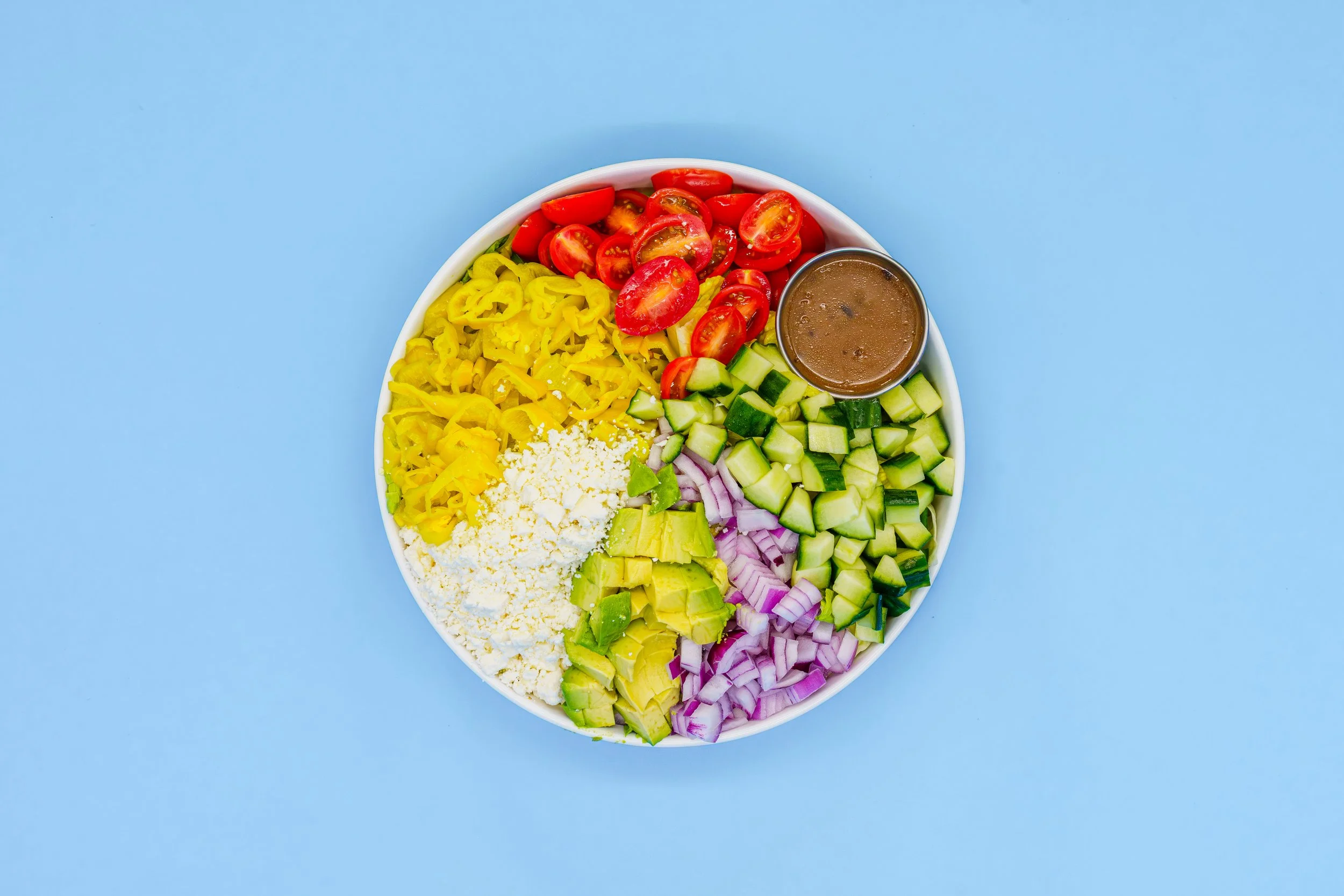 A white oval dish containing cherry tomatoes, sliced yellow bell peppers, cucumber cubes, chopped red onions, avocado pieces, shredded cheese, and a small cup of brown dressing on a light blue background.