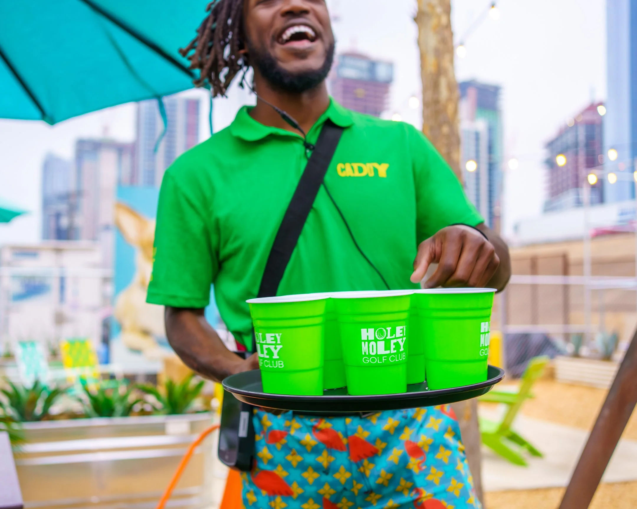 A person with dreadlocks wearing a green shirt and colorful shorts holding a tray with four green cups that have white text reading "Hole-in-One Golf Club." The person appears to be outside in front of a city skyline with high-rise buildings, trees, and other people.