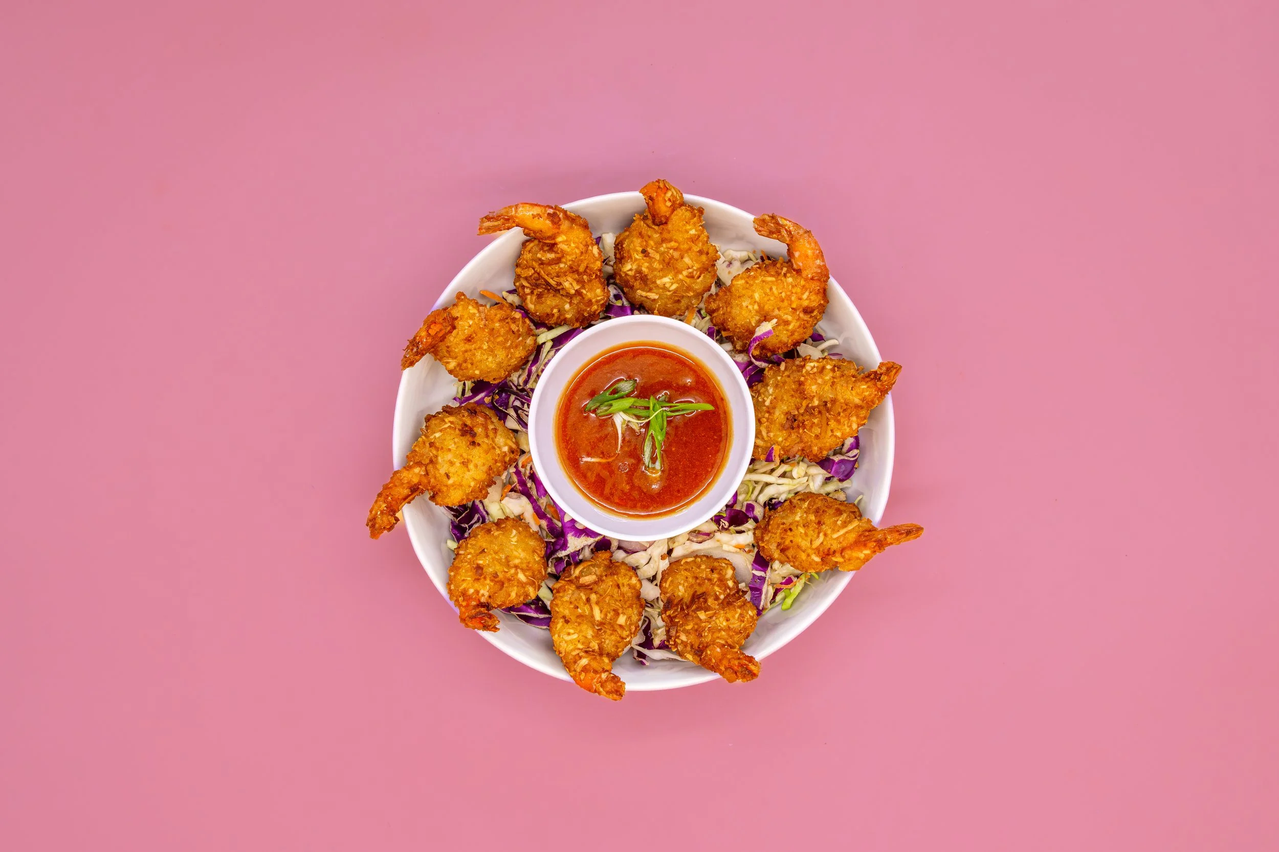 A white bowl of fried shrimp with breaded coating, served with a small cup of red dipping sauce garnished with green onion, on a bed of shredded purple and white cabbage, against a pink background.