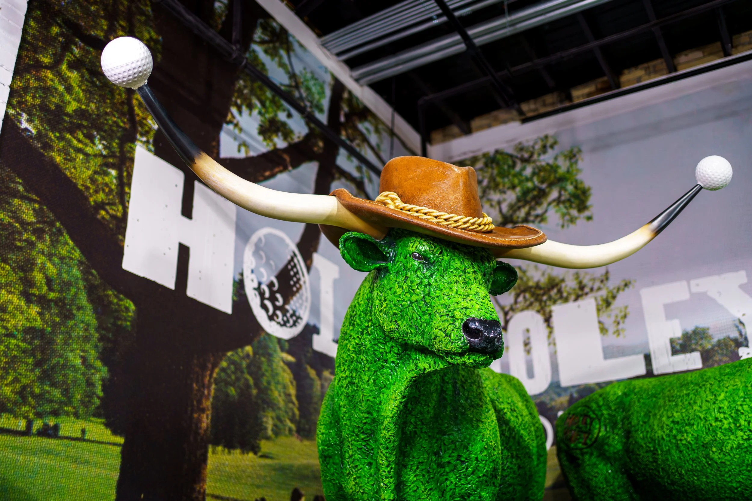 A green, textured bear statue wearing a brown cowboy hat with a gold rope band, and with long, white bullhorns with golf ball tips extending from its head.