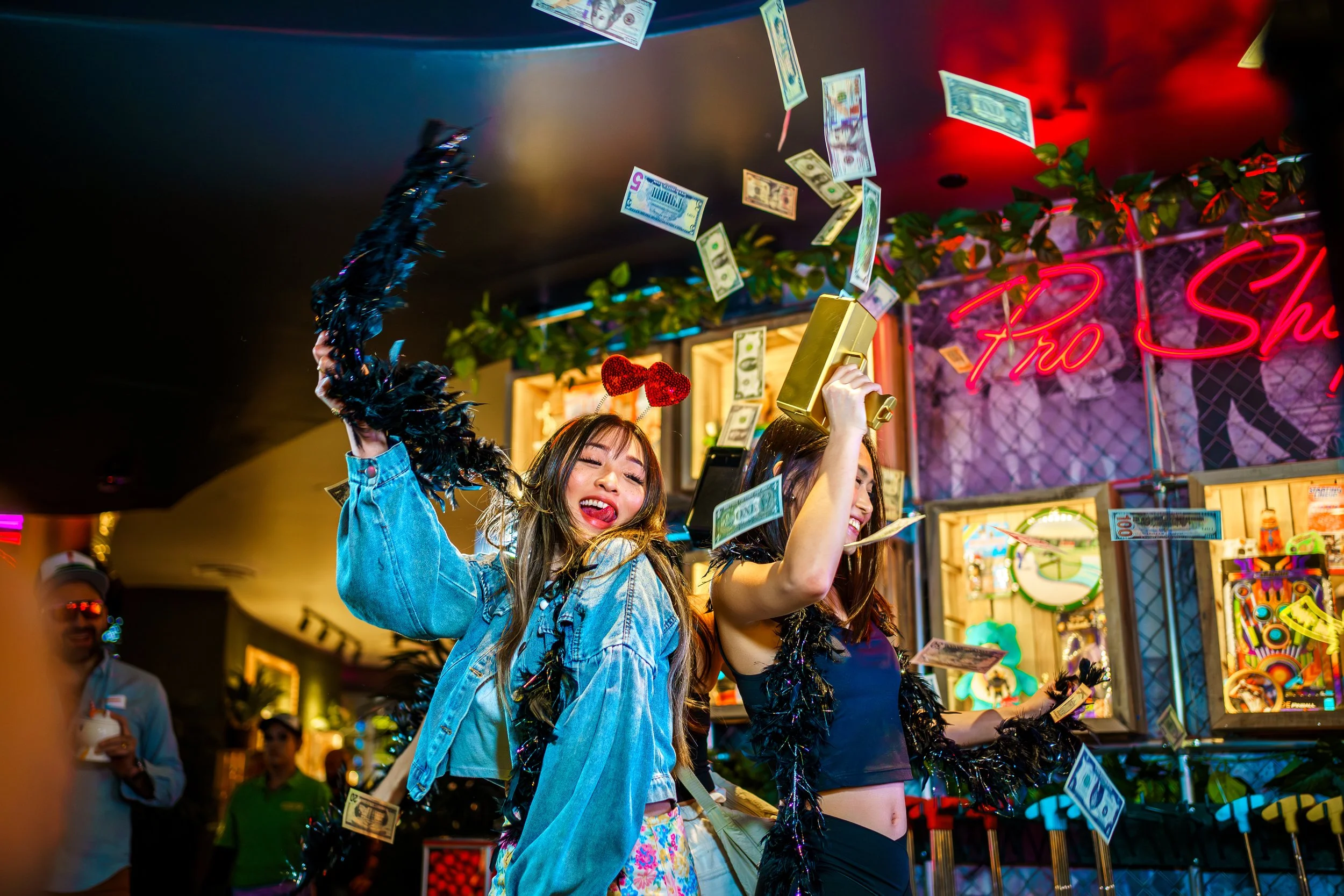Two young women celebrate at a carnival game booth with falling dollar bills, holding novelty gold and red trophies, with neon signs and arcade prizes in the background.