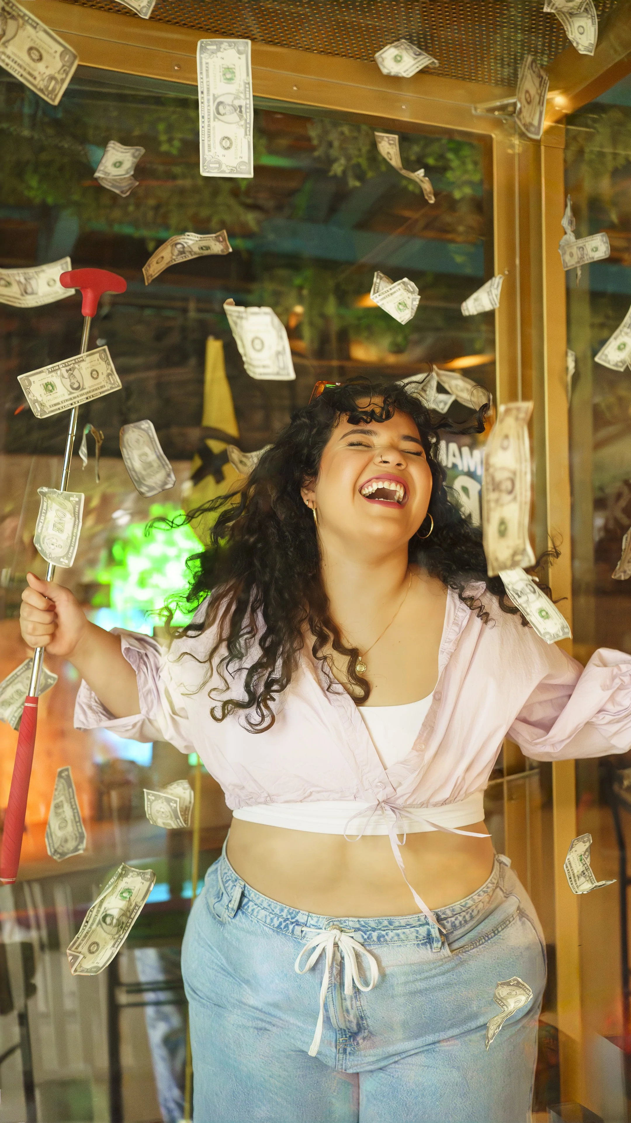 A young woman laughing and celebrating inside a glass booth with dollar bills floating around her, holding a hook with a red handle.