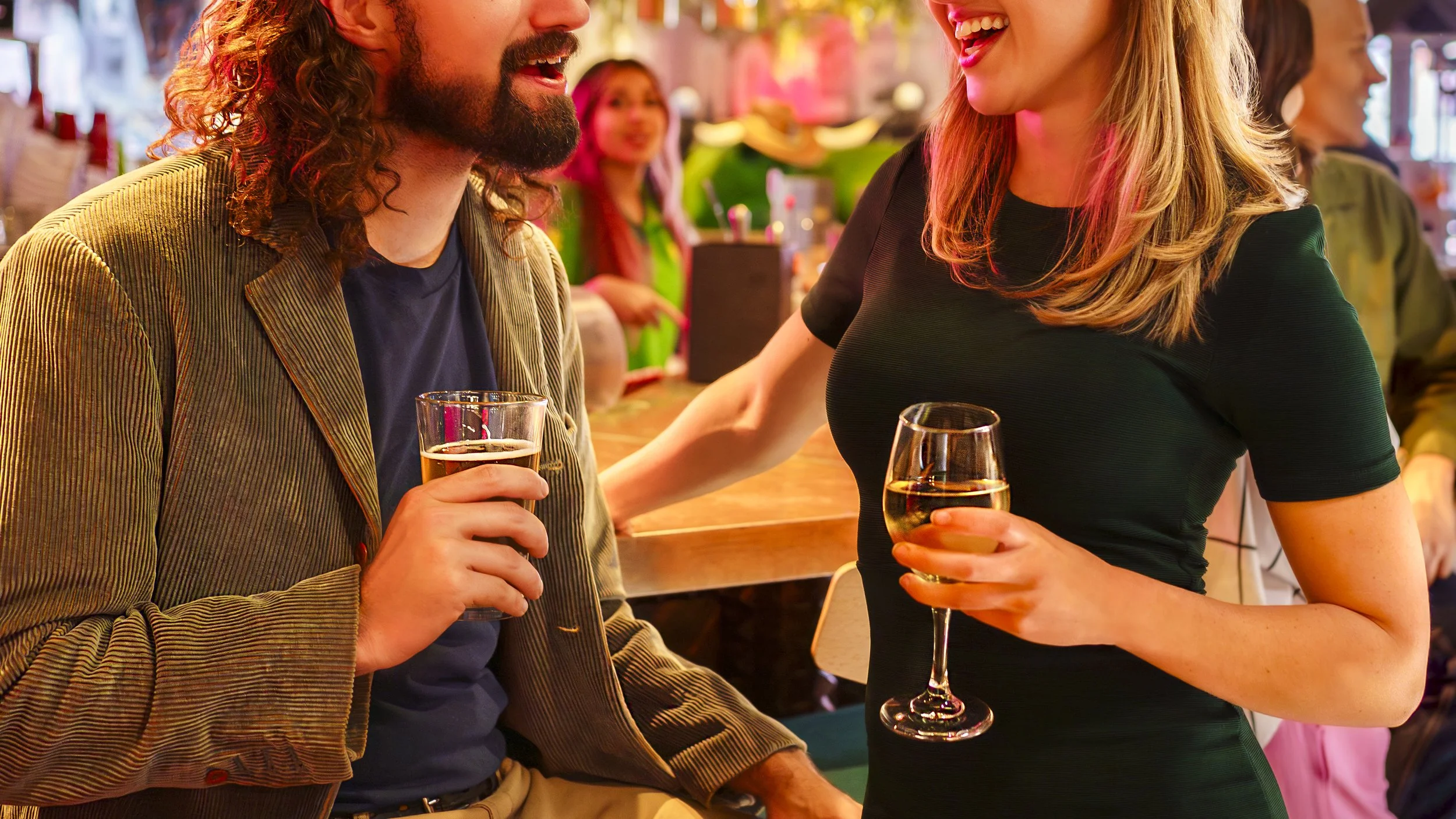 Two people enjoying drinks and conversing at a lively social event. The man has long, curly hair and a beard, holding a glass of beer; the woman in a black dress with blonde hair is holding a glass of white wine, smiling.