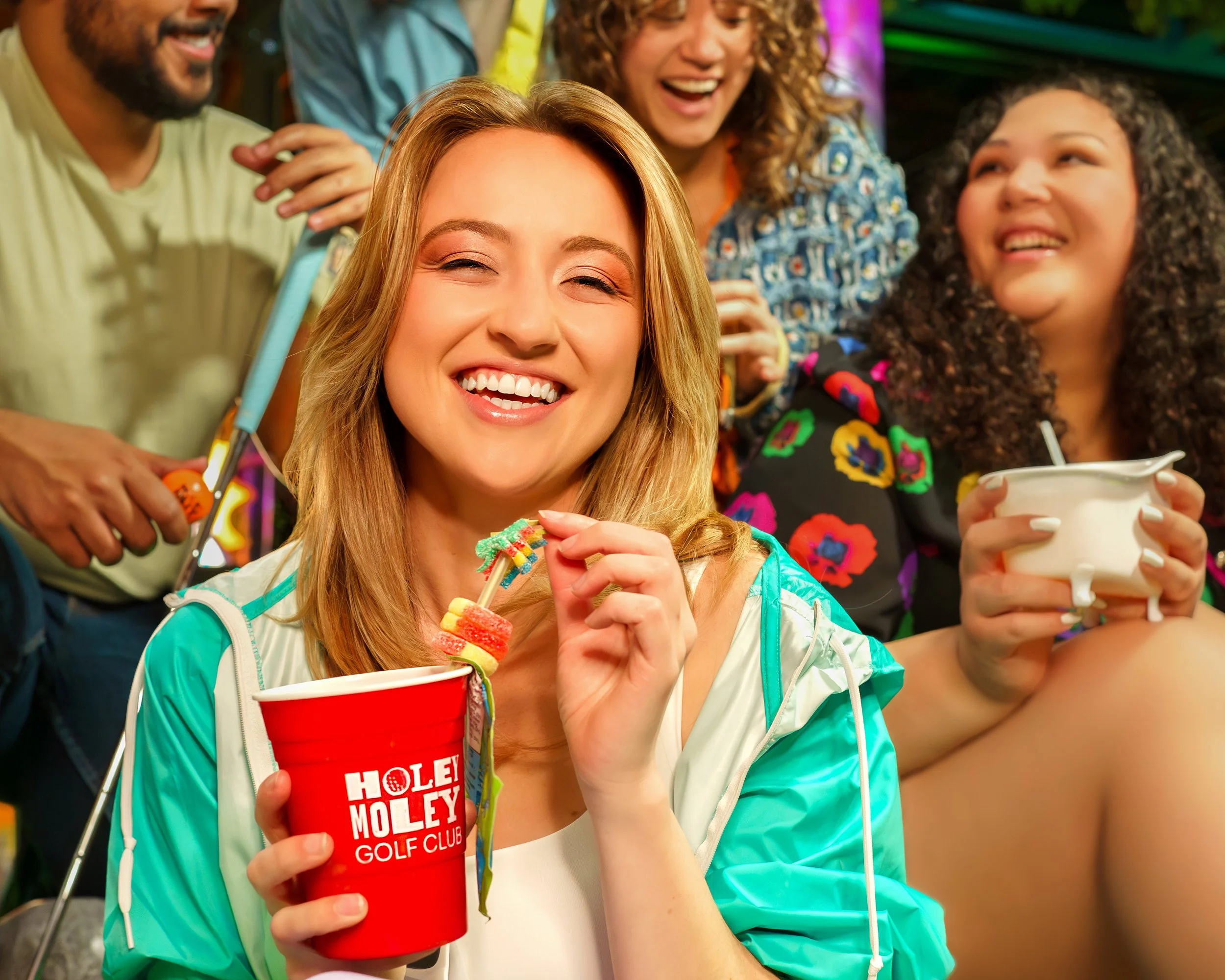 Group of people at a lively social gathering, smiling and enjoying drinks and food, with colorful background lighting.
