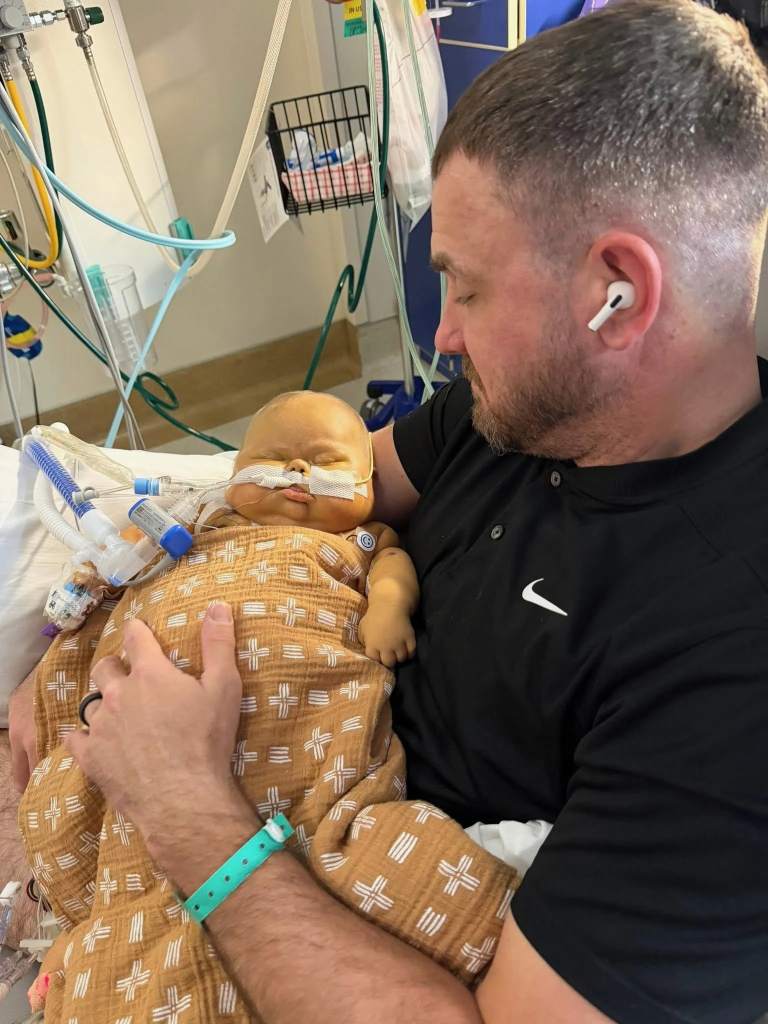 A man with short hair and a black Nike shirt holding a premature baby in a hospital. The baby is connected to medical equipment, wearing a patterned outfit, and appears to be sleeping. The man is wearing a teal hospital wristband.