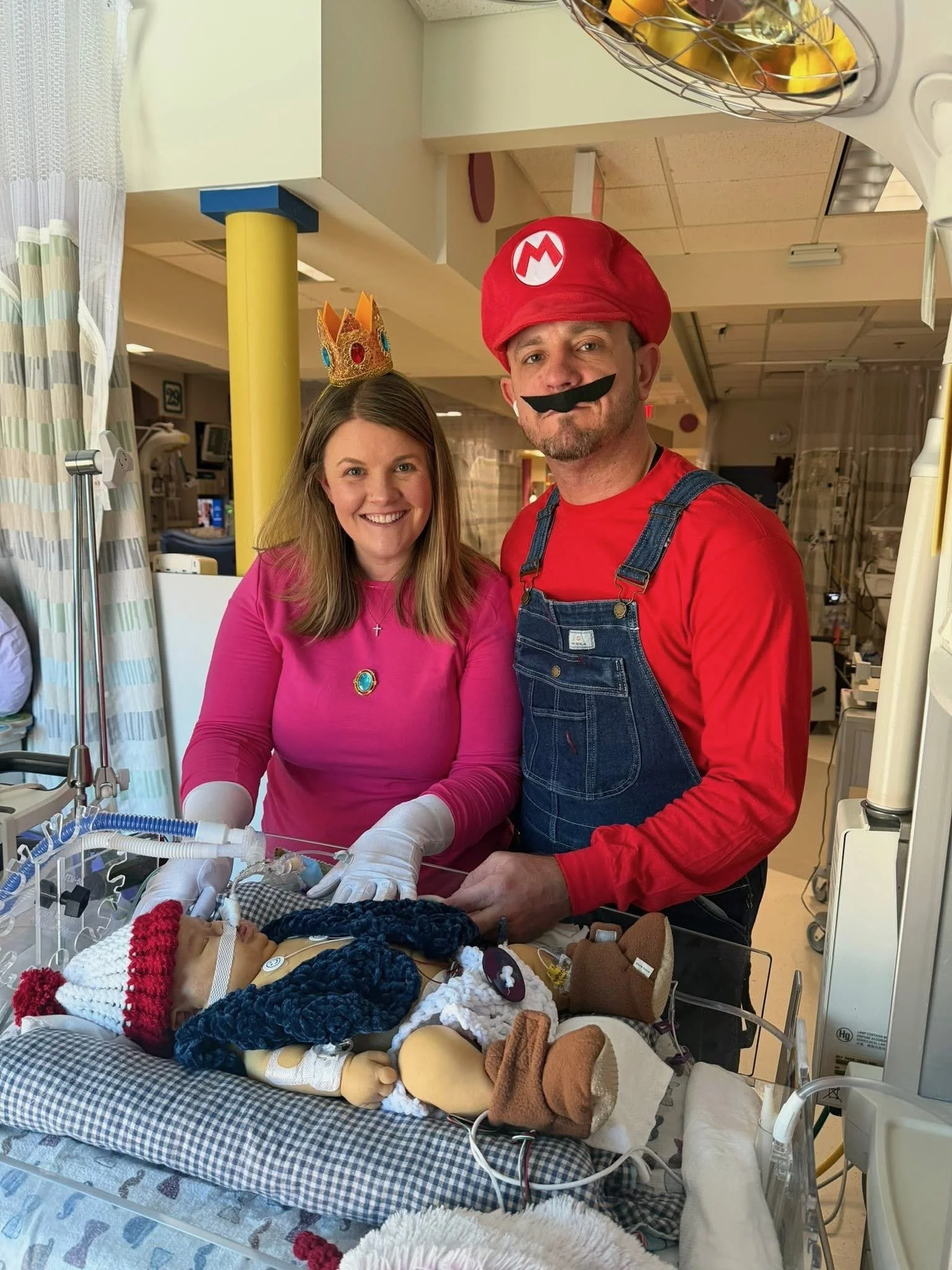 A woman and a man dressed as Mario and Princess Peach from Super Mario in a hospital room, standing next to a hospital bassinet filled with knitted toys and stuffed animals for a newborn baby.