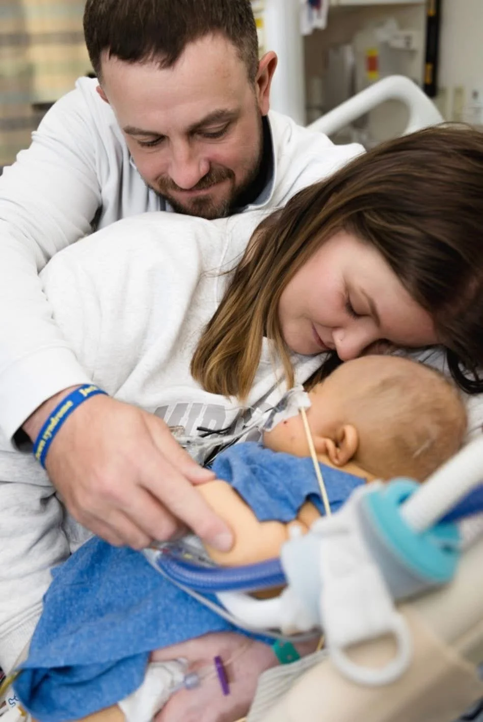 A couple holding a sleeping infant in a hospital room, with the father smiling and the mother kissing the baby's forehead.