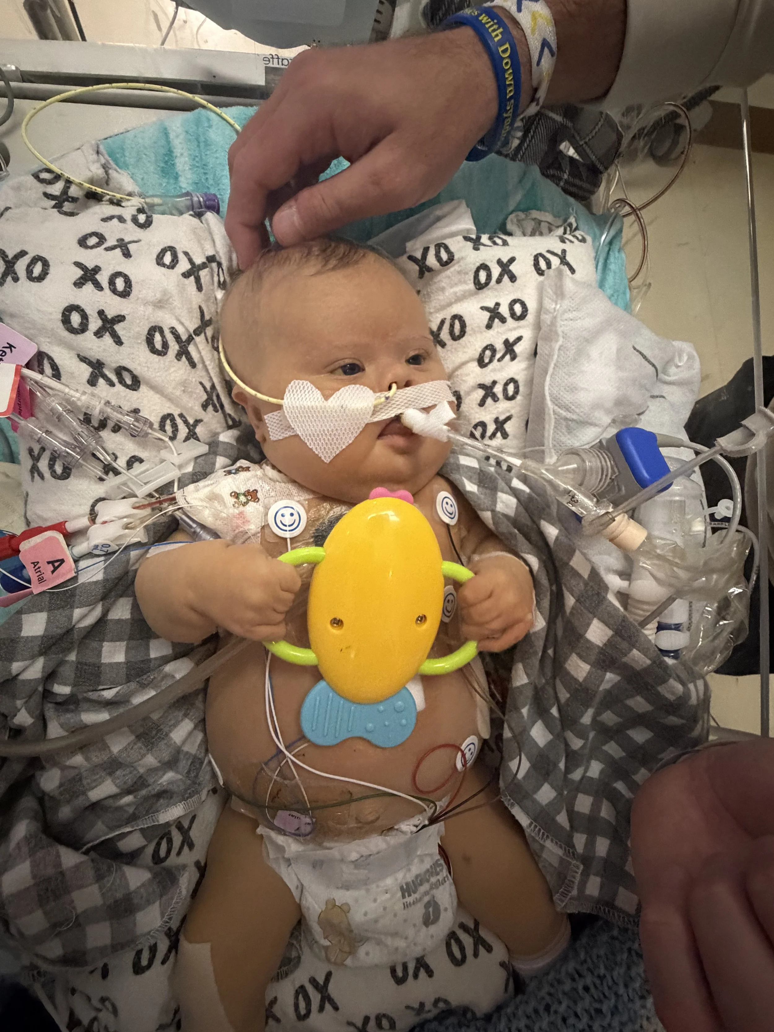 A young child in a hospital bed with medical tubes, monitors, and a pacifier, holding a yellow toy, with another person beside them
