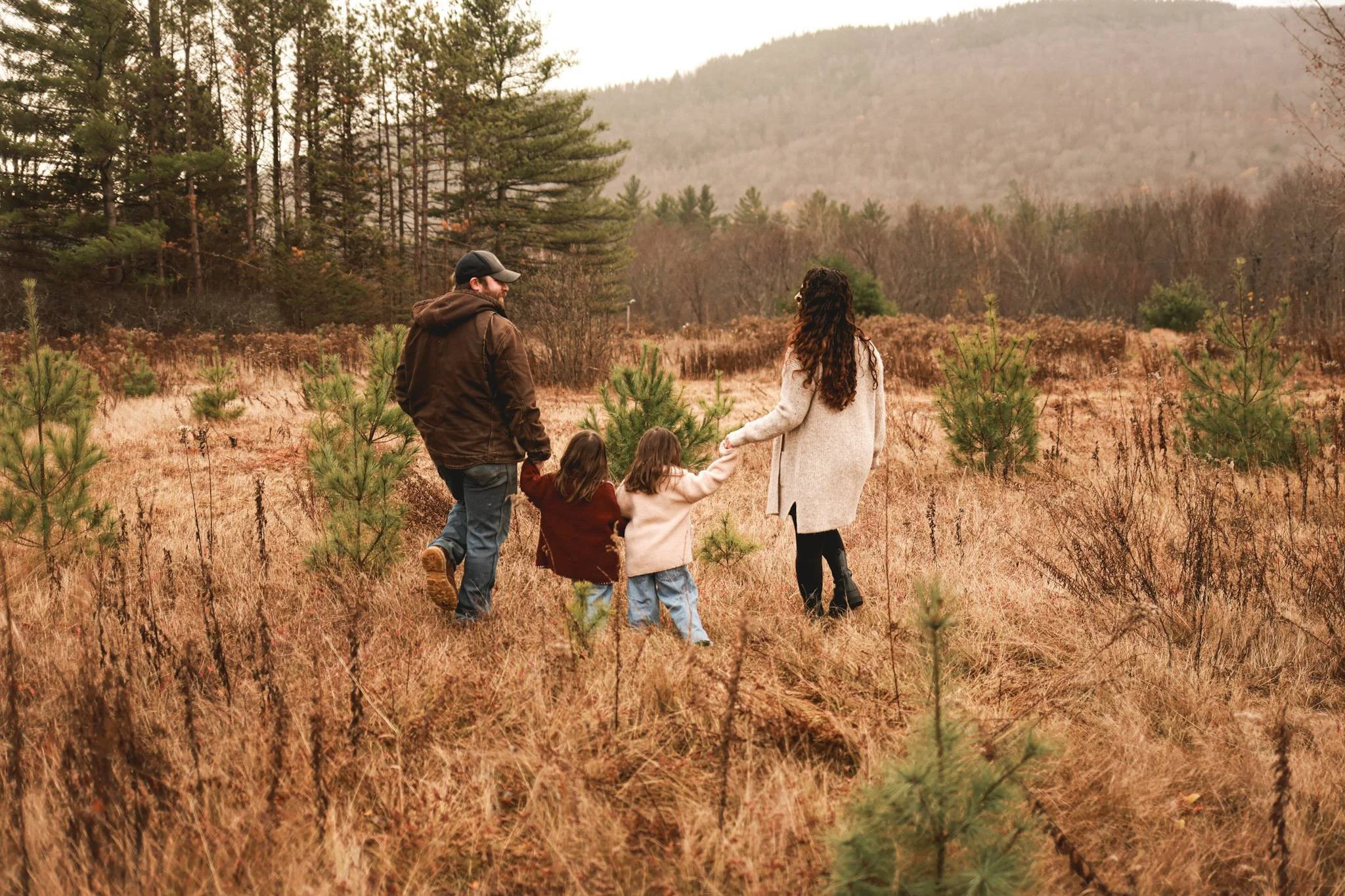 A family of five walking through a field with dry grass and small pine trees, surrounded by a forest and mountains in the background during autumn.