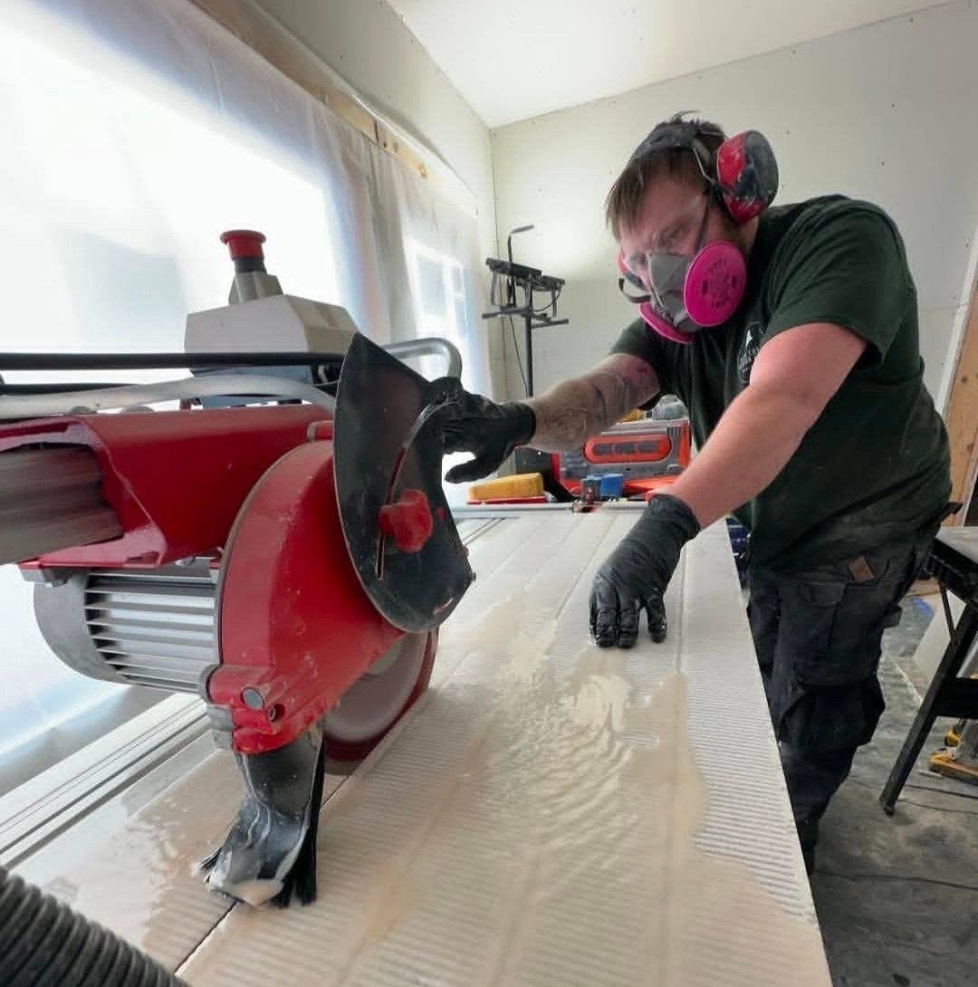 Man using a red circular saw while wearing safety gear including gloves, ear protection, and a mask in a workshop.
